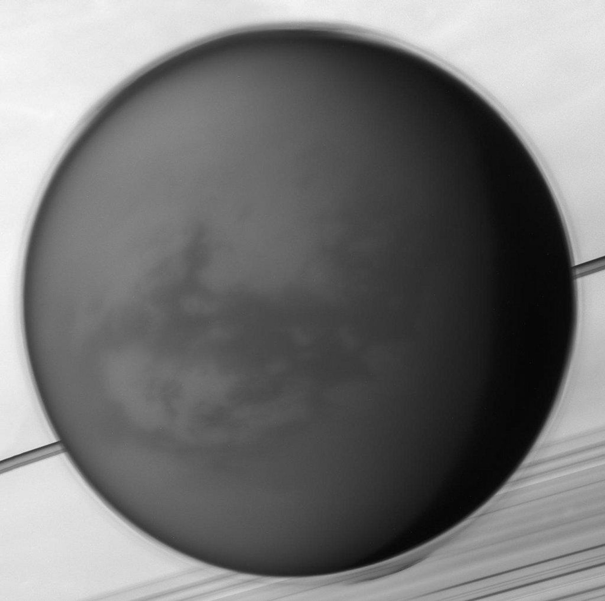 A black and white image of Saturn's largest moon, Titan, taken by the Cassini spacecraft. The image shows Titan's anti-Saturn side, the side always facing away from the ringed gas giant. Titan's dense atmosphere and haze are visible, along with the dark dune-filled region known as Shangri-La.