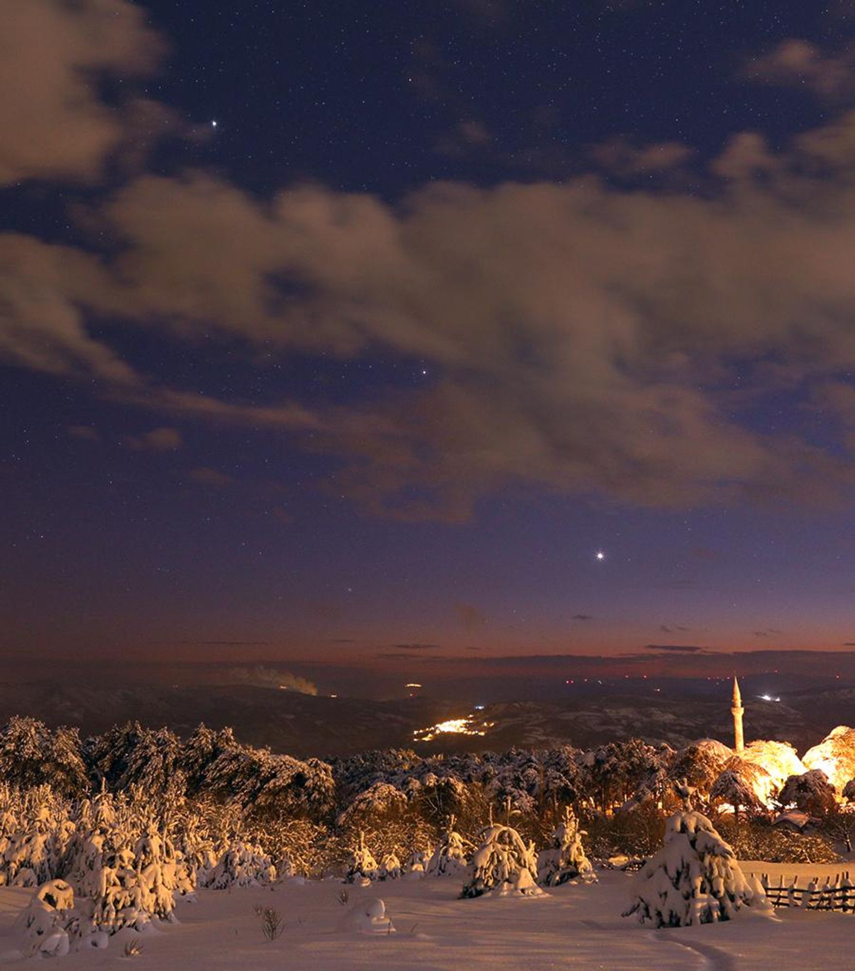 A breathtaking view of the December solstice evening sky from Kirazli, Turkey, showcasing a celestial alignment featuring Venus, Saturn, Jupiter, and Comet Leonard. The brightest celestial beacon, Venus, shines near the southwestern horizon, while Saturn glimmers between clouds, and Jupiter dominates the sky above. Comet Leonard, the fainter celestial visitor, forms a nearly equilateral triangle with Venus and Saturn. This picturesque scene is framed by a snow-covered landscape, adding to its wintery charm.