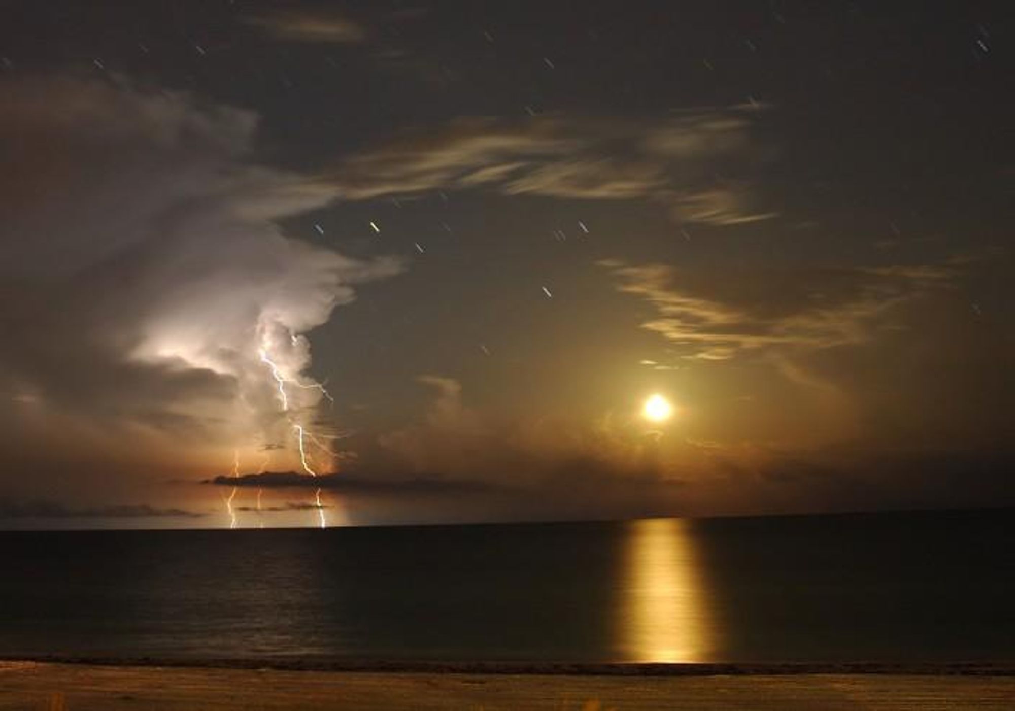 A long exposure photograph of a moonset over the Gulf of Mexico. The moon is setting in the distance, while lightning strikes in the foreground. The clouds are illuminated by the moon and the lightning, and the stars are visible as streaks in the sky. The bright yellowish star trail just above and right of the lightning flash is red giant star Antares.