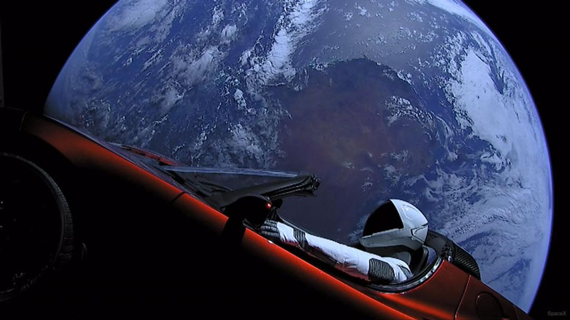 A red Tesla roadster with a spacesuit-clad mannequin, dubbed Starman, at the wheel, orbits the Earth in a photo taken from one of three cameras mounted on the car.  The Earth is seen in the background, with clouds and land masses visible.  The car is on its way to the asteroid belt and will eventually orbit the Sun.