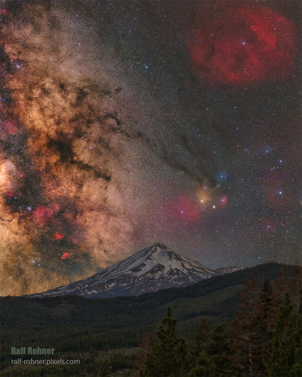 A composite image of Mount Shasta, California, with the Milky Way Galaxy arching over the snow-capped peak. The vibrant colors of the Rho Ophiuchi cloud complex and the red emission nebula surrounding the star Zeta Ophiuchi are visible above the mountain, along with the bright orange star Antares. The image was taken during the blue hour and later that night with a two-panel panorama tracking the background sky.