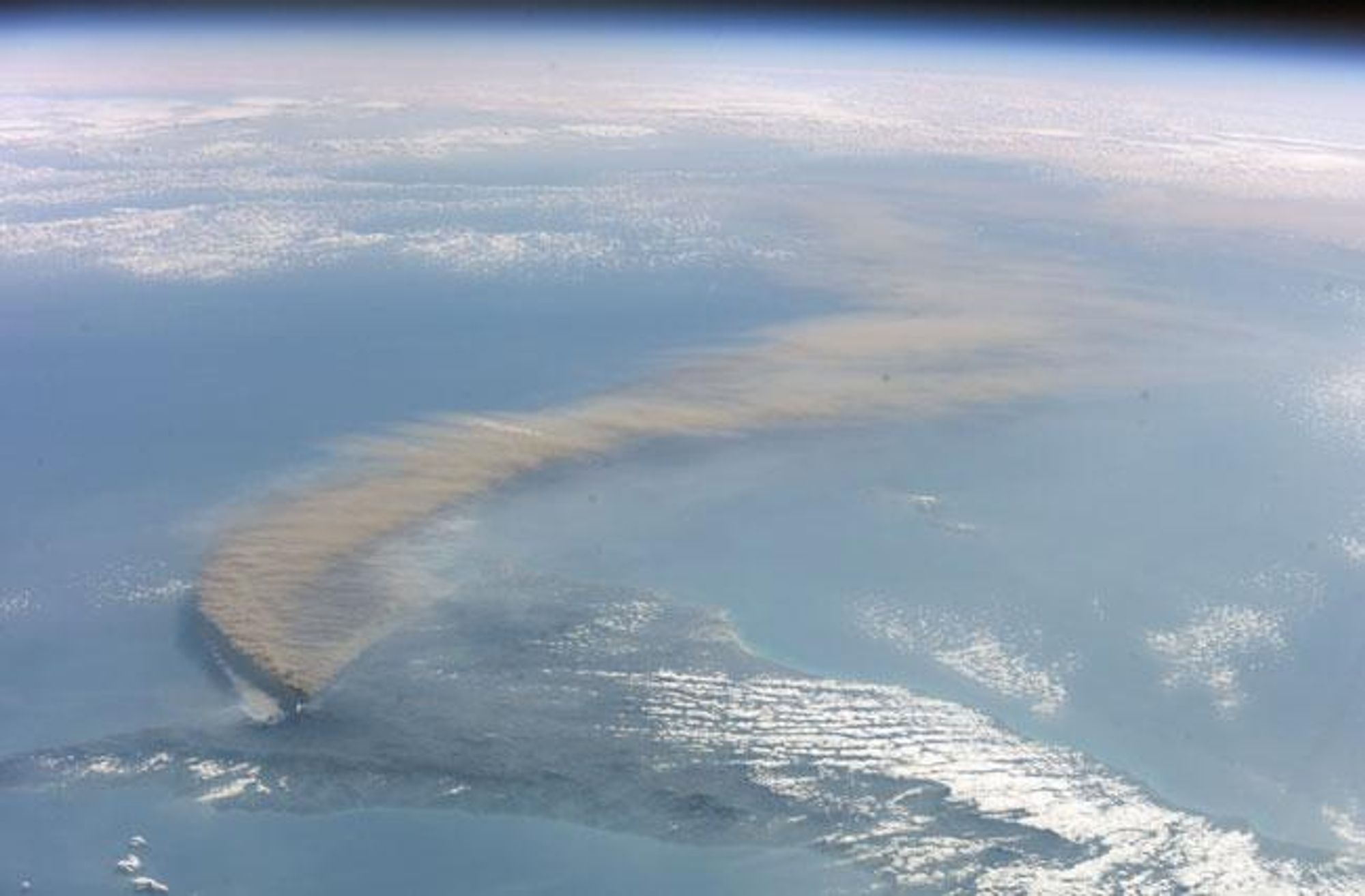 An astronaut aboard the International Space Station captured this image of a plume of ash from Mount Etna. The ash is visible as a thin, light-colored band stretching across the image.  Light colored smoke to the left of the ash plume is due to forest fires started by lava flow on the volcano's north face. This view is looking towards the southeast.