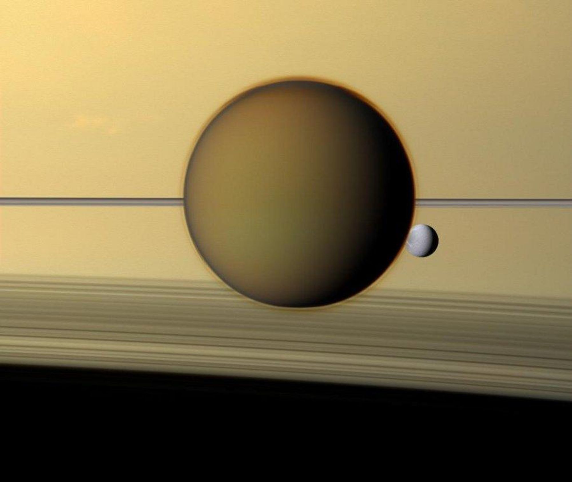 A close-up image of Saturn, captured by the Cassini spacecraft, showcasing Titan and Dione with the rings in the foreground. Titan, the largest moon of Saturn, appears as a large, hazy sphere with Dione, the third largest moon, visible through its atmosphere. The rings cast arcing shadows across Saturn's cloud tops at the bottom of the frame.