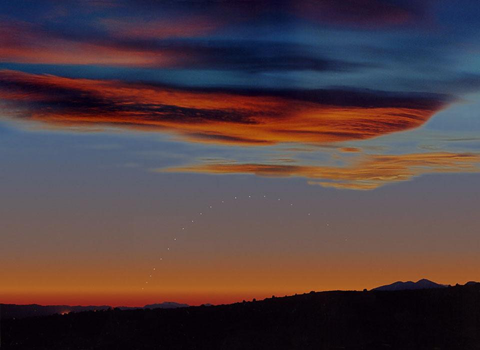 A composite image of Mercury's position in the sky over a sunset in Spain. The image was created by taking several photos of Mercury at different times during March 2000 and superimposing them on a single photogenic sunset. Each photo was taken when the Sun was 10 degrees below the horizon. Mercury can be seen as a line of white dots in the sky, moving in an arc.