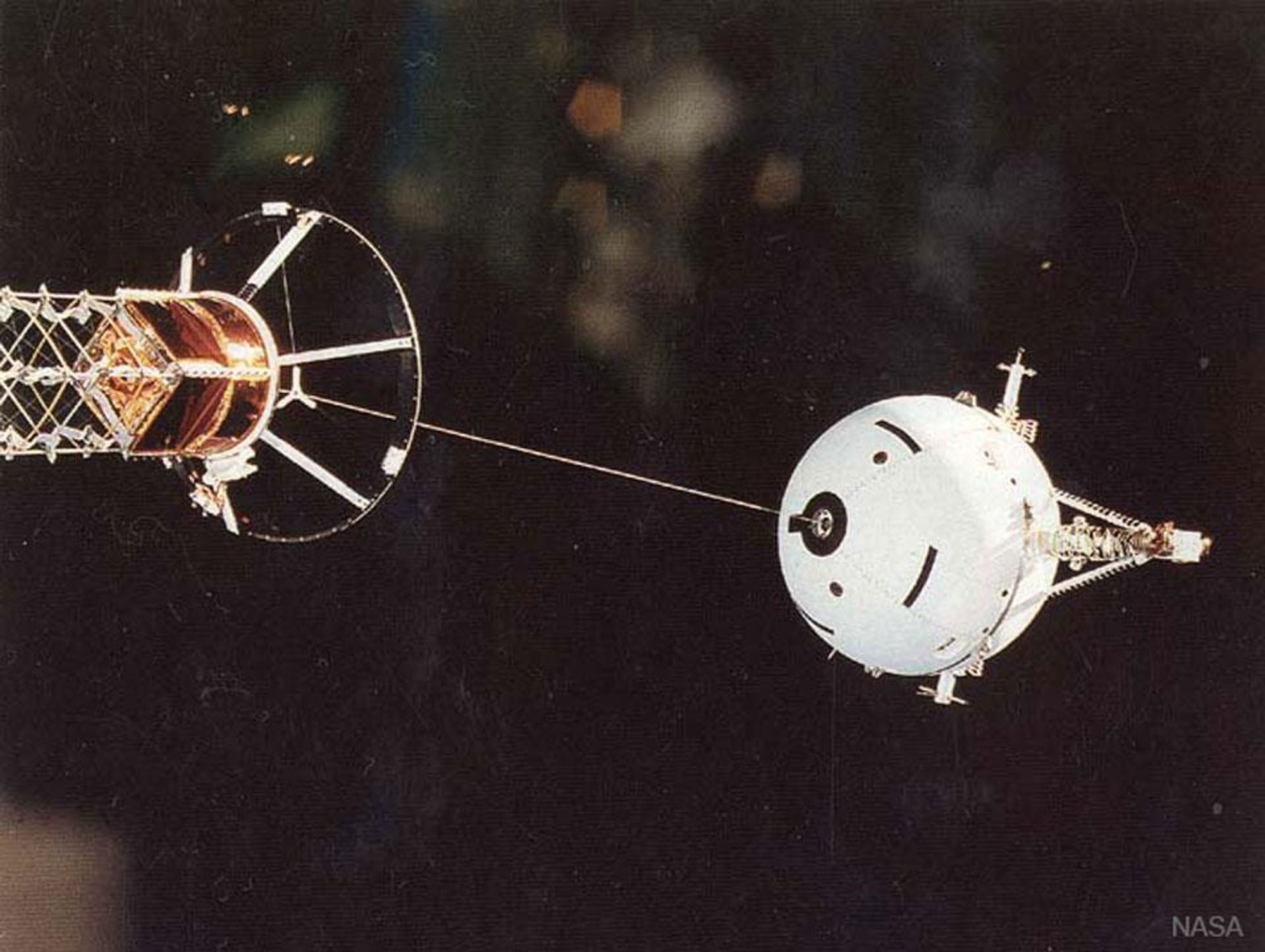 A black and white photo of the Tethered Satellite System 1 (TSS-1) being deployed by the Space Shuttle Atlantis in 1992. TSS-1 was a revolutionary attempt at using a long tether to stabilize satellites, generate electricity, and allow for easy transportation. Though TSS-1 failed to live up to its promise, many valuable lessons were learned.