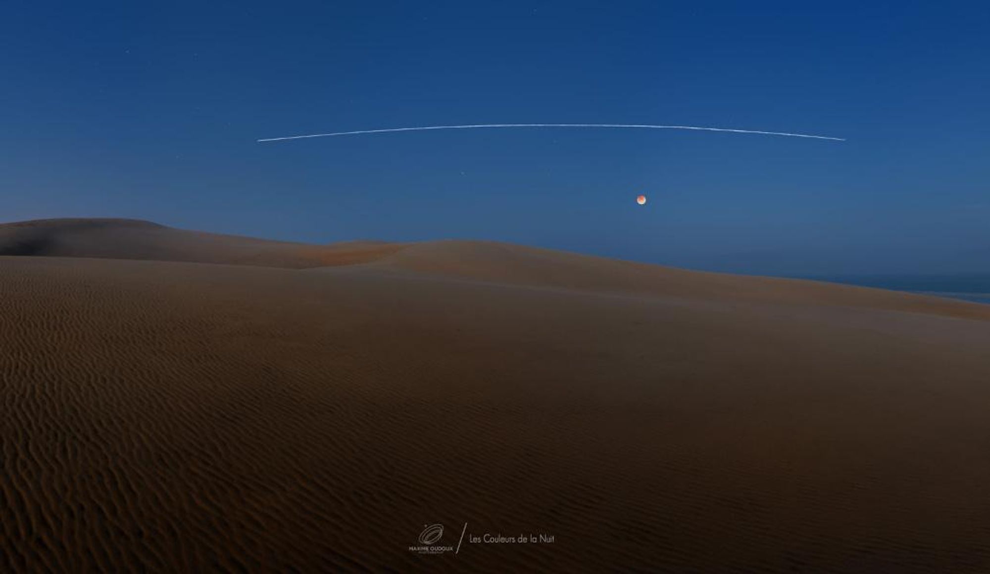 A panoramic view of the Dune of Pilat in France during a total lunar eclipse on May 16. The moon is in the upper right corner of the image, partially obscured by the earth's shadow. The International Space Station is visible as a bright, flat arc across the sky. The image was captured over 5 minutes using a series of consecutive images.