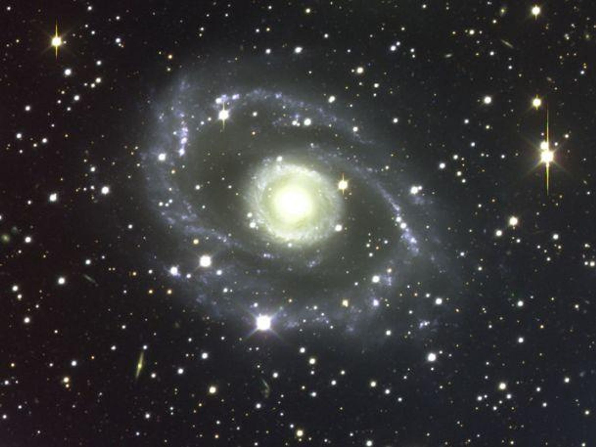 A stunning photograph of a spiral galaxy, ESO 269-57, captured by the Very Large Telescope in Chile. The galaxy is seen face-on and is about 150 million light-years away, and 200,000 light-years across. The brighter foreground stars are marked by diffraction spikes caused by the telescope and yellow vertical stripes due to saturated digital camera pixels. The galaxy is located in the constellation Centaurus, which is one of the most striking constellations in the southern sky.  The image also shows tantalizing wisps of more distant, faint galaxies in the background.