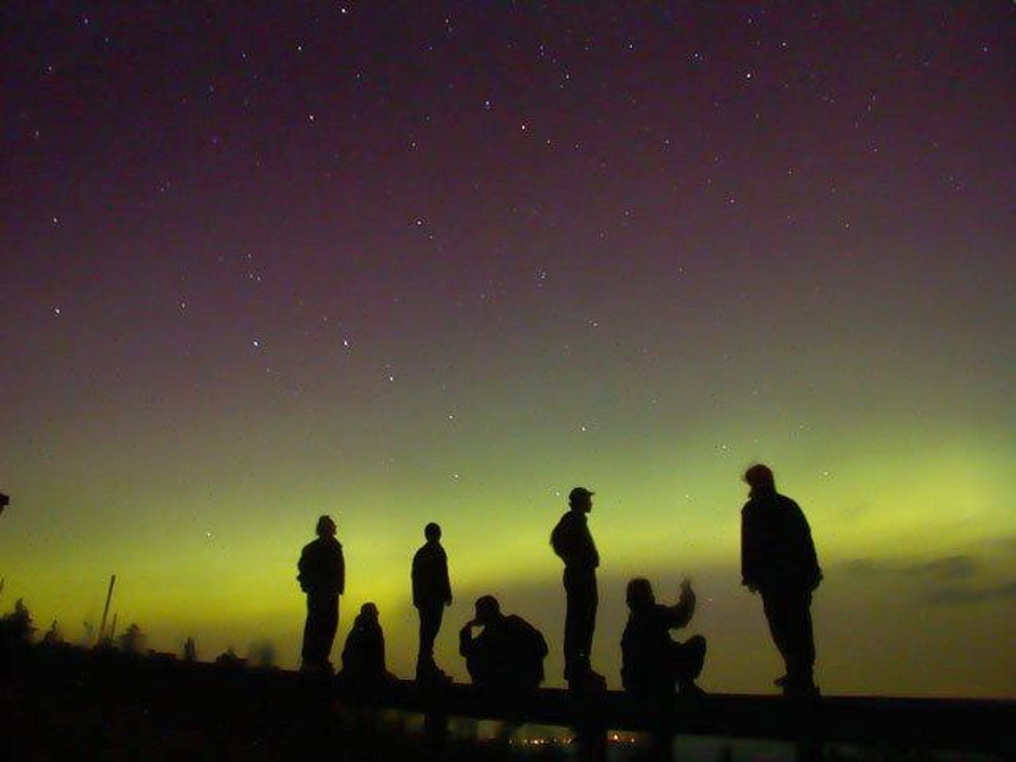 A group of silhouetted people stand and gaze up at the night sky, which is alive with green aurora borealis, the milky way galaxy, stars, and the rising moon. They are gazing up at the Perseid meteor shower which should be visible for the rest of the week.