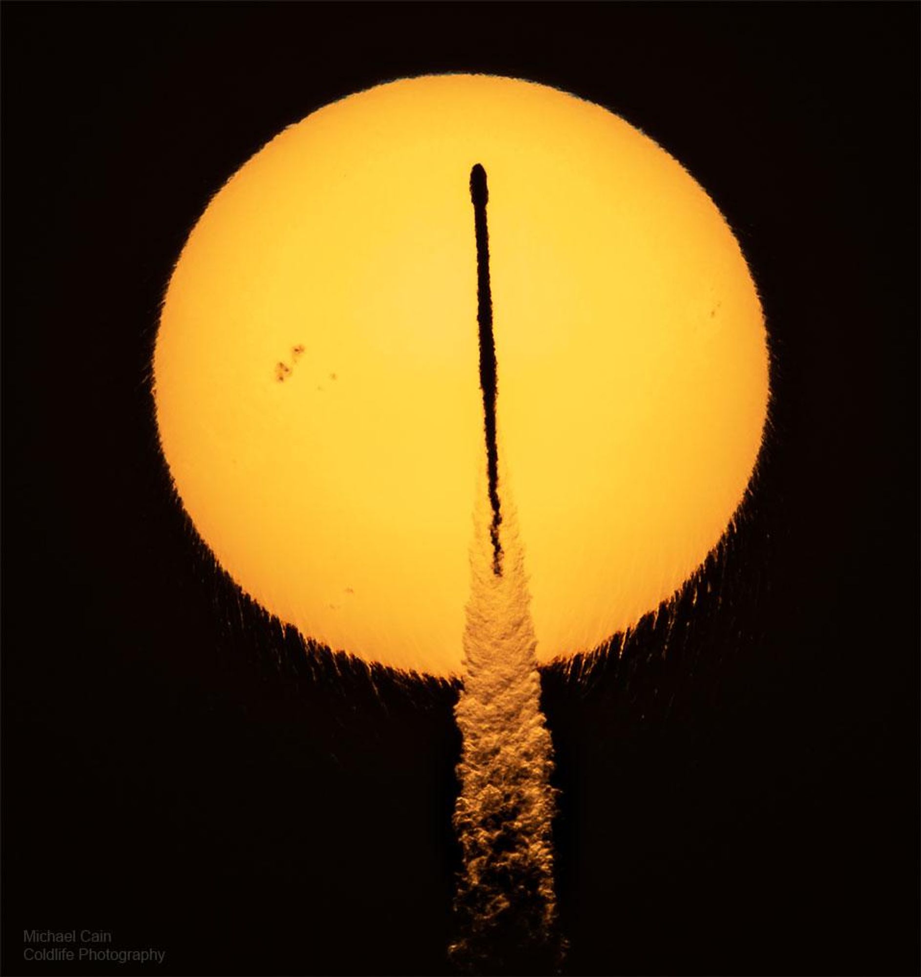 A SpaceX Falcon 9 rocket launches at sunrise, casting a silhouette against the sun. The rocket's exhaust plume glows brightly, and the sun's lower edge shows peculiar drip-like ripples due to refraction. An active sunspot region can be seen in the upper left of the image.