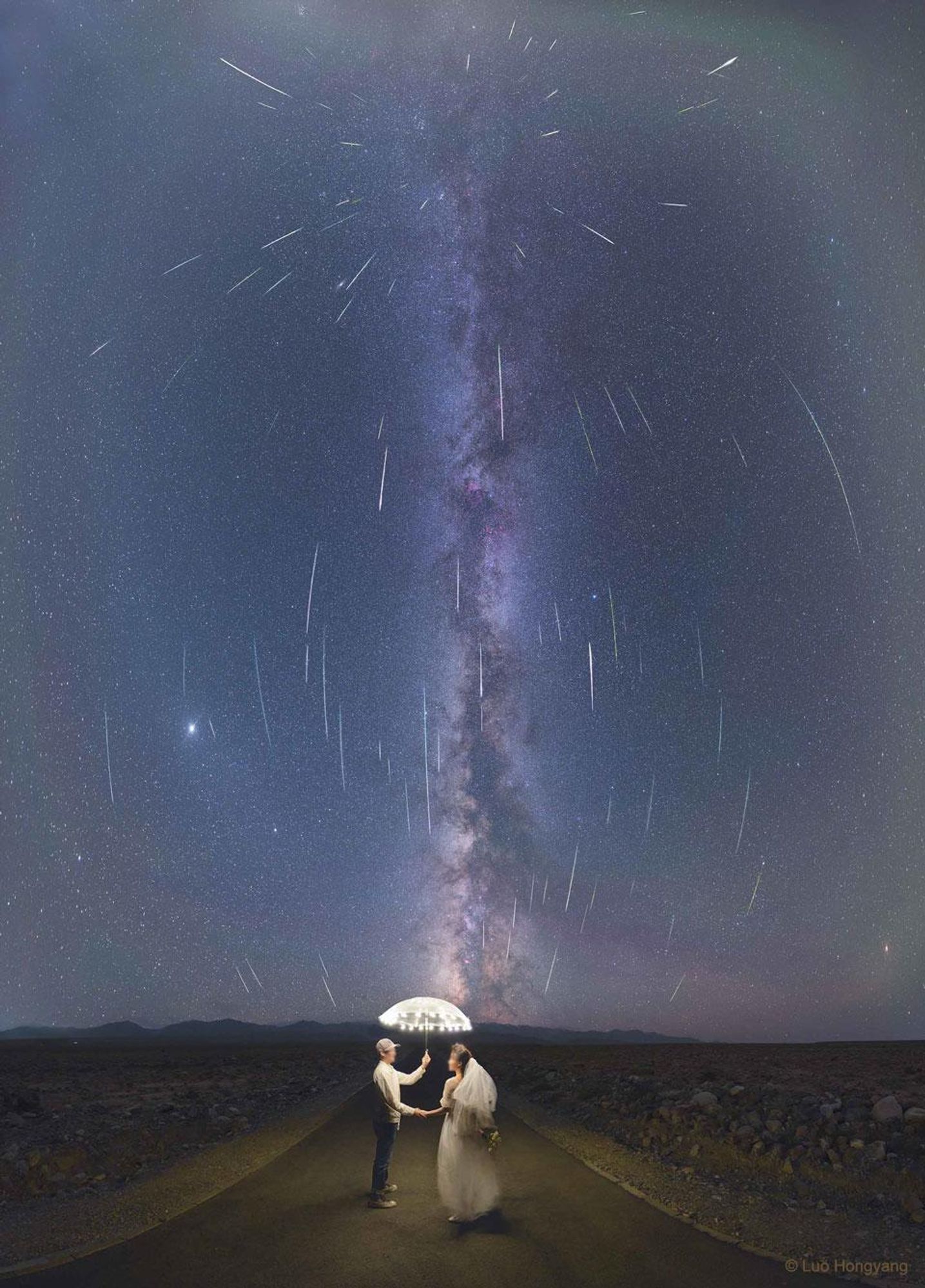 A composite image of the Perseid meteor shower over Jiuquan City, Gansu Province, China, featuring a couple standing under an umbrella on a road with the Milky Way Galaxy and the planets Jupiter and Saturn in the background. The image captures the curvature of the sky and the radiant point of the meteor shower in the constellation Perseus.