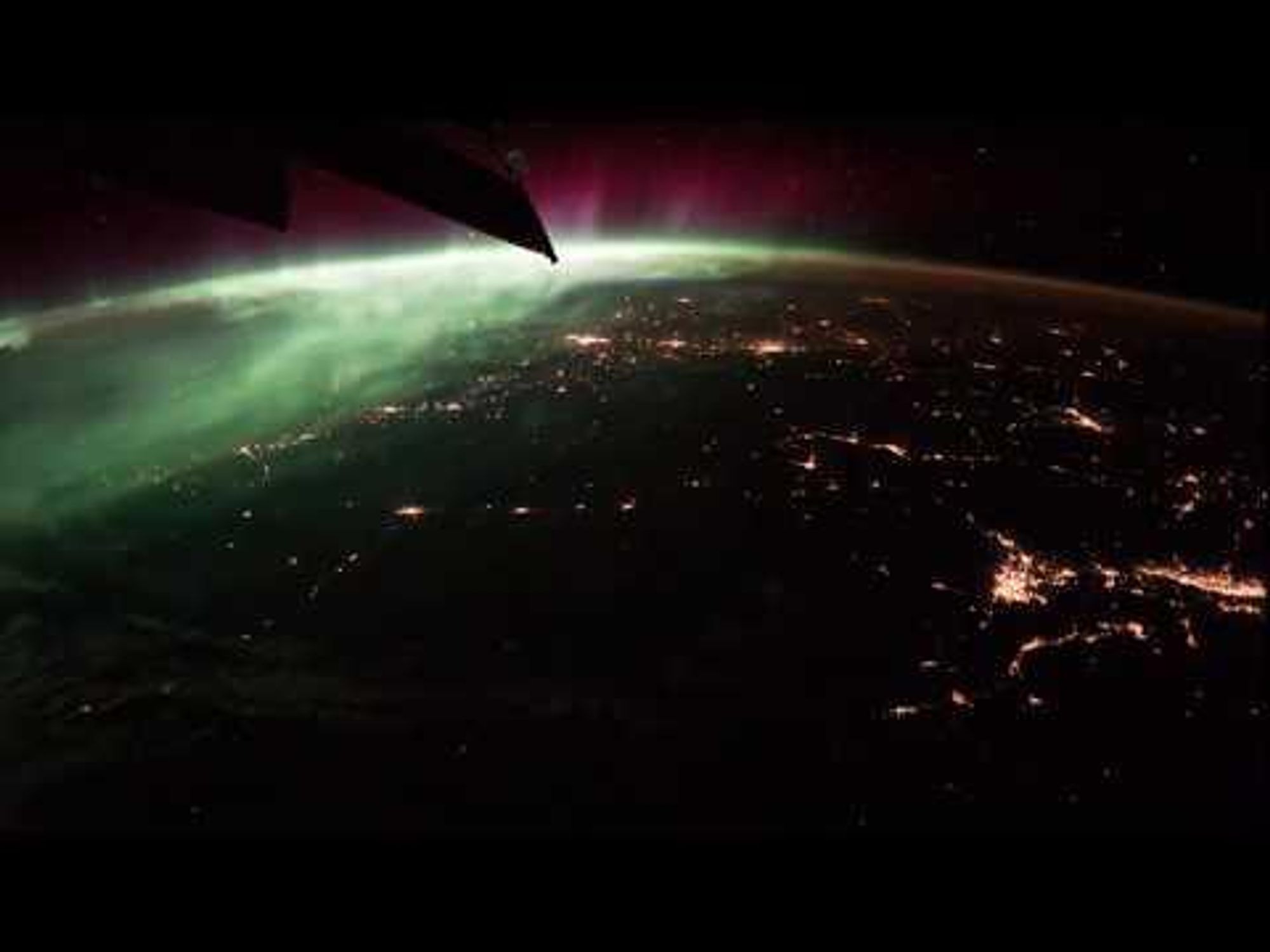 A view of Earth from space, showing the International Space Station (ISS) orbiting over North America and Europe, with green and red auroras in the upper left corner, city lights below, and lightning strikes flashing in the distance. Stars can be seen rising through the greenish-gold glow of Earth's atmosphere.