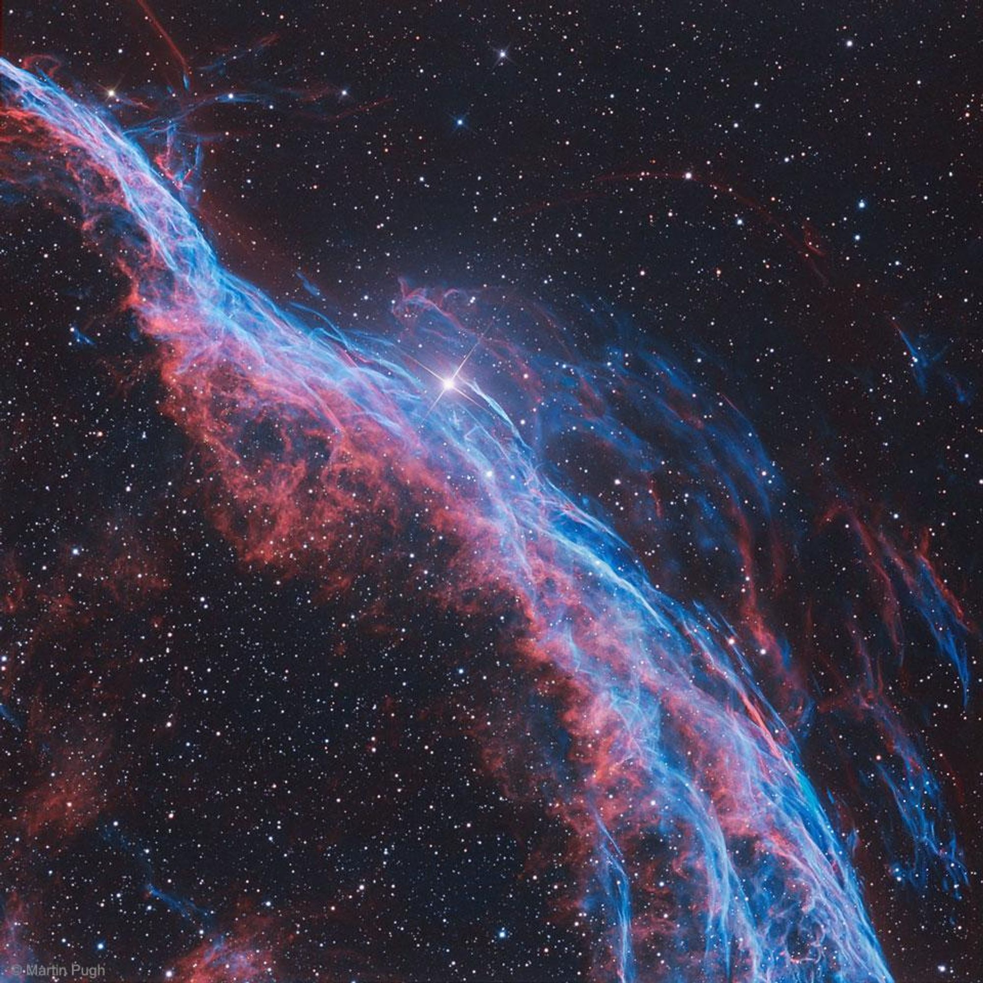 A stunning telescopic image of the Witch's Broom Nebula (NGC 6960), a western segment of the Veil Nebula, a supernova remnant. The image shows the glowing filaments of the nebula in red and blue-green, representing atomic hydrogen and oxygen gas, respectively. The nebula is located about 1400 light-years away towards the constellation Cygnus and spans about 35 light-years. A bright star, 52 Cygni, can be seen in the frame, but is unrelated to the supernova remnant.