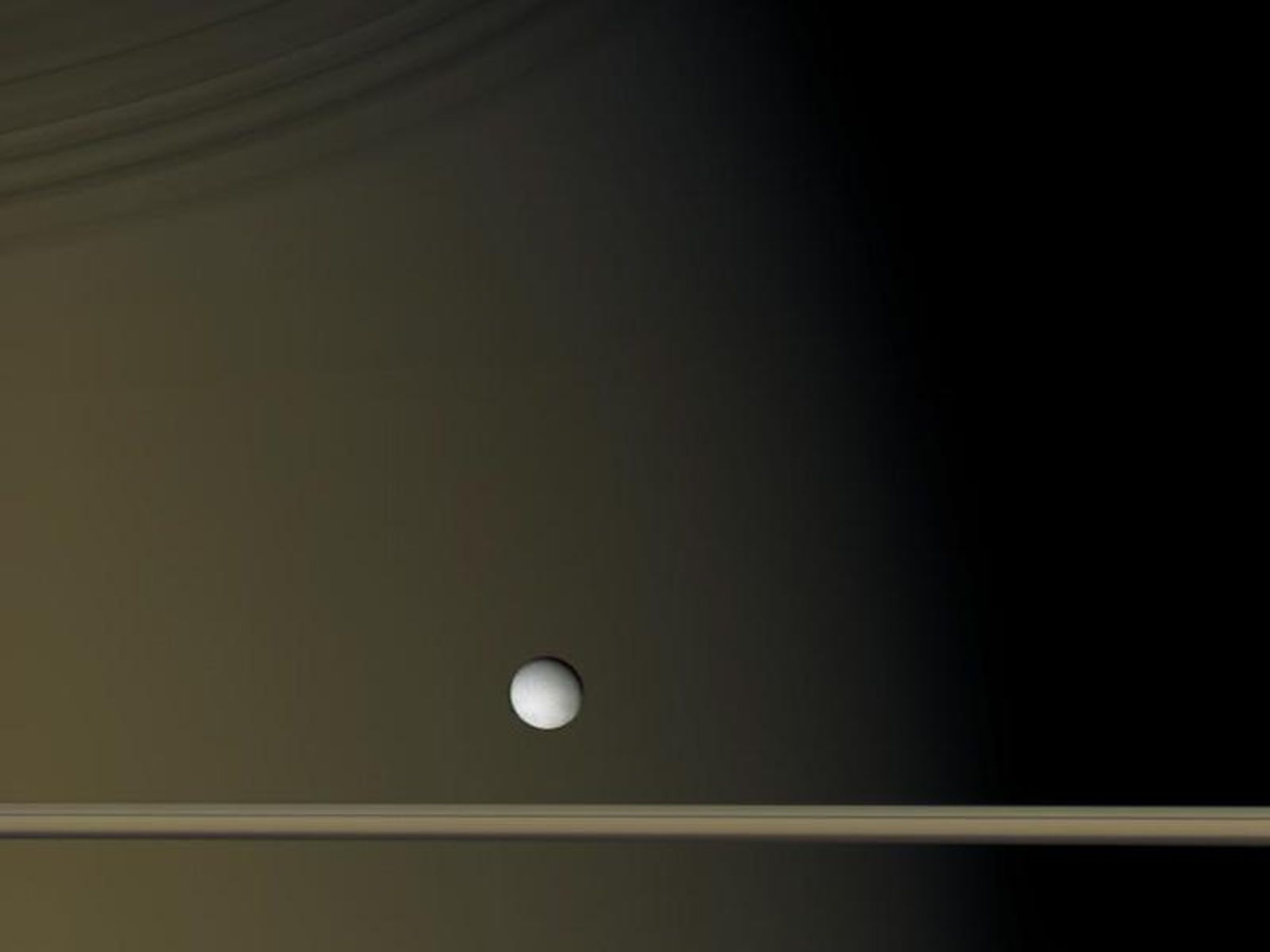 A surreal image of Saturn, its rings, and its moon Enceladus, captured by the Cassini spacecraft.  Enceladus appears white against the backdrop of Saturn's rings, which are seen edge-on and appear golden brown due to their dirt content.  The planet Saturn is visible in the background, with its terminator (the line between night and day) visible across its face.