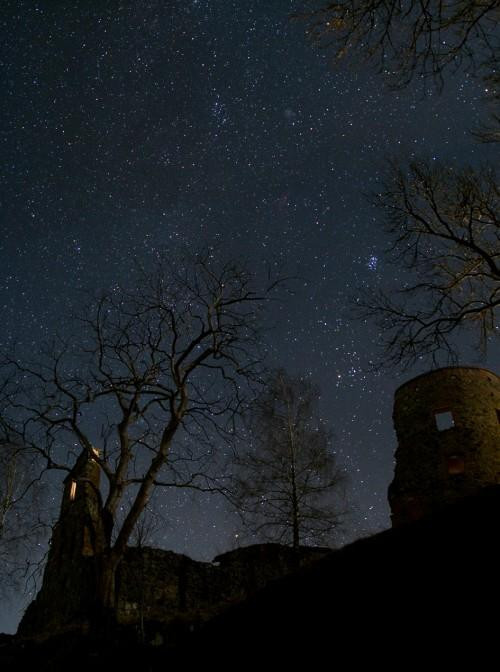 A silhouetted medieval castle tower stands against a dark night sky filled with stars, including the Pleiades star cluster, Aldebaran, and Comet Holmes. The castle, known as Mörby Castle, is located in Sweden near Lake Skedviken.