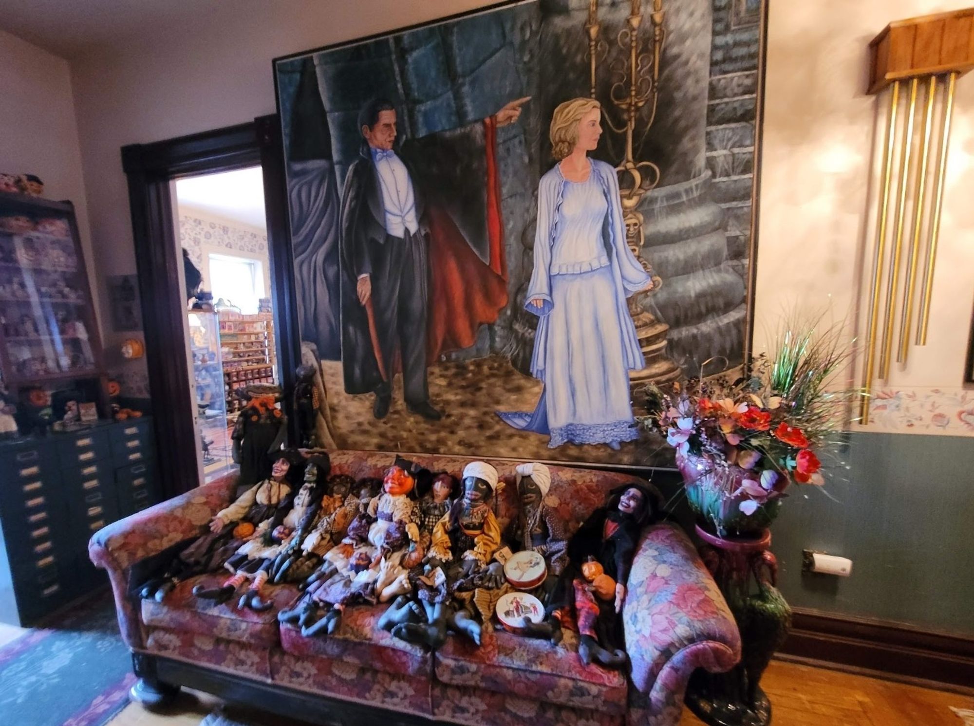 A large painting of a caped vampire pointing off into the distance, while a young woman in a blue dress stands next to him and looks to where he's pointing. In front of the painting is an old couch with a ton of little creepy dolls.