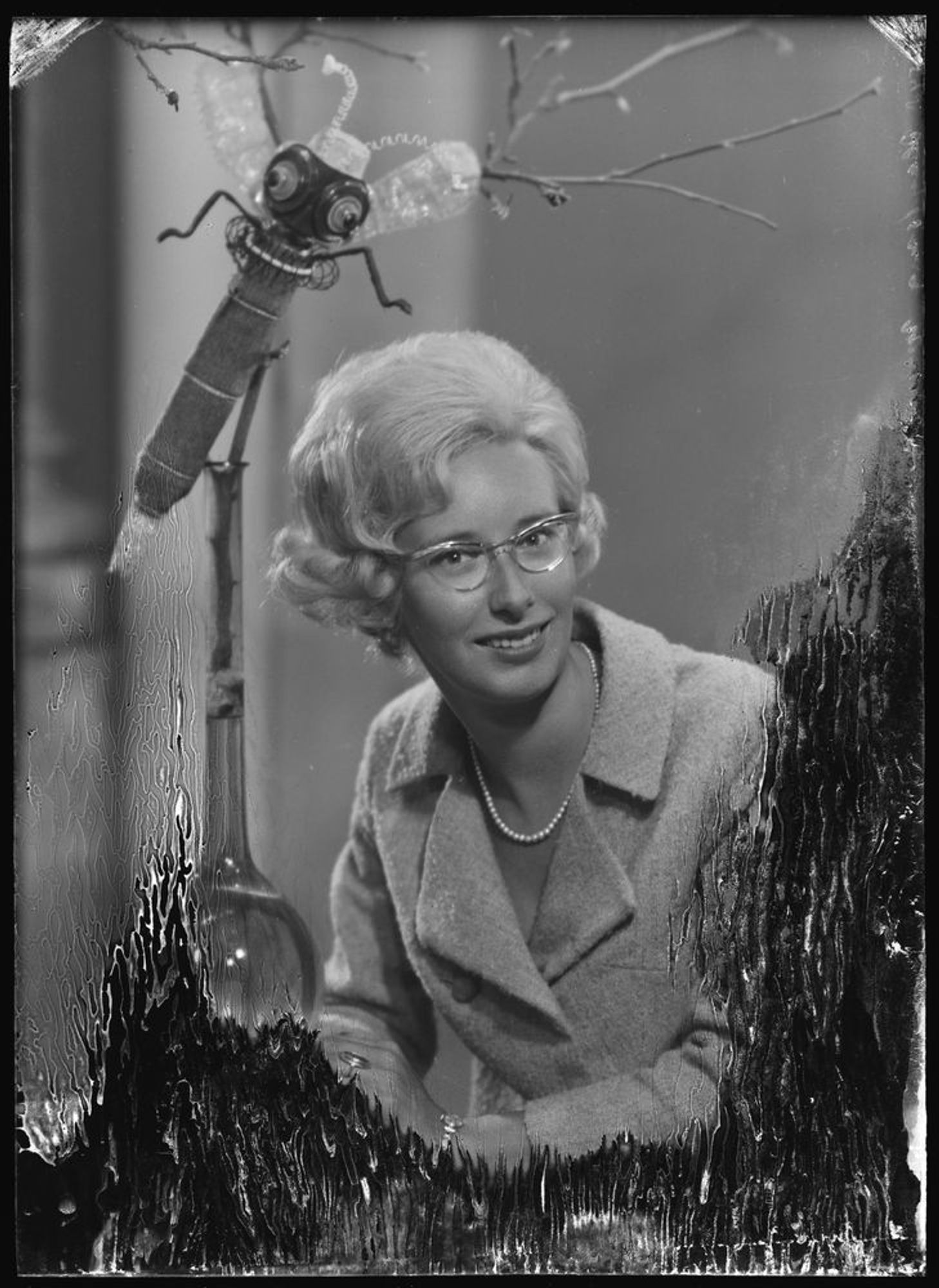 portrait of a smiling blond woman in a suit jacket and glasses and pearls, sitting down with arms folded in front of her. Next to her elbow is some kind of glass tube with a bulbous bottom and a long stem, Attached to the stem is a cute, quirky, handmade-looking insect with wings and antennae and a little pillbox hat, mayb? And only two legs. And a stinger, ooh she's packin