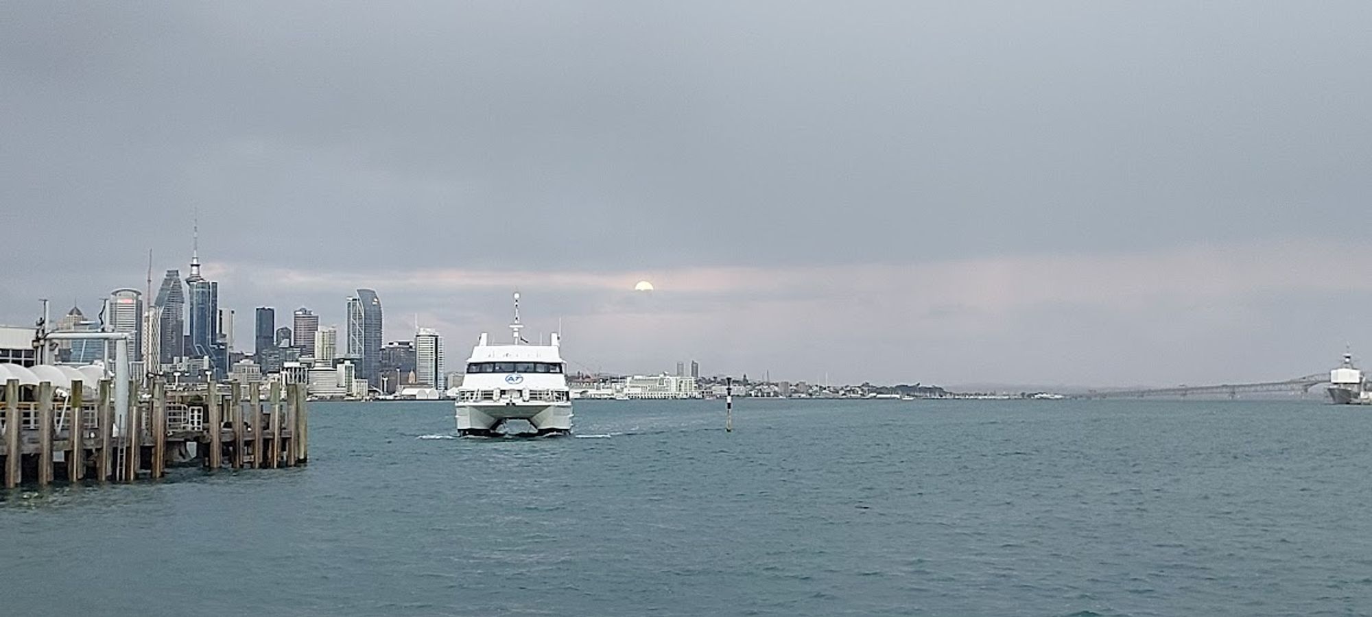 Auckland harbour with a pier on the left and the skyline in the background. A ferry is coming towards the viewer. In the grey and cloudy sky is what looks like a sun setting. However, if you know Auckland, you'll recognise this is actually the moon setting.