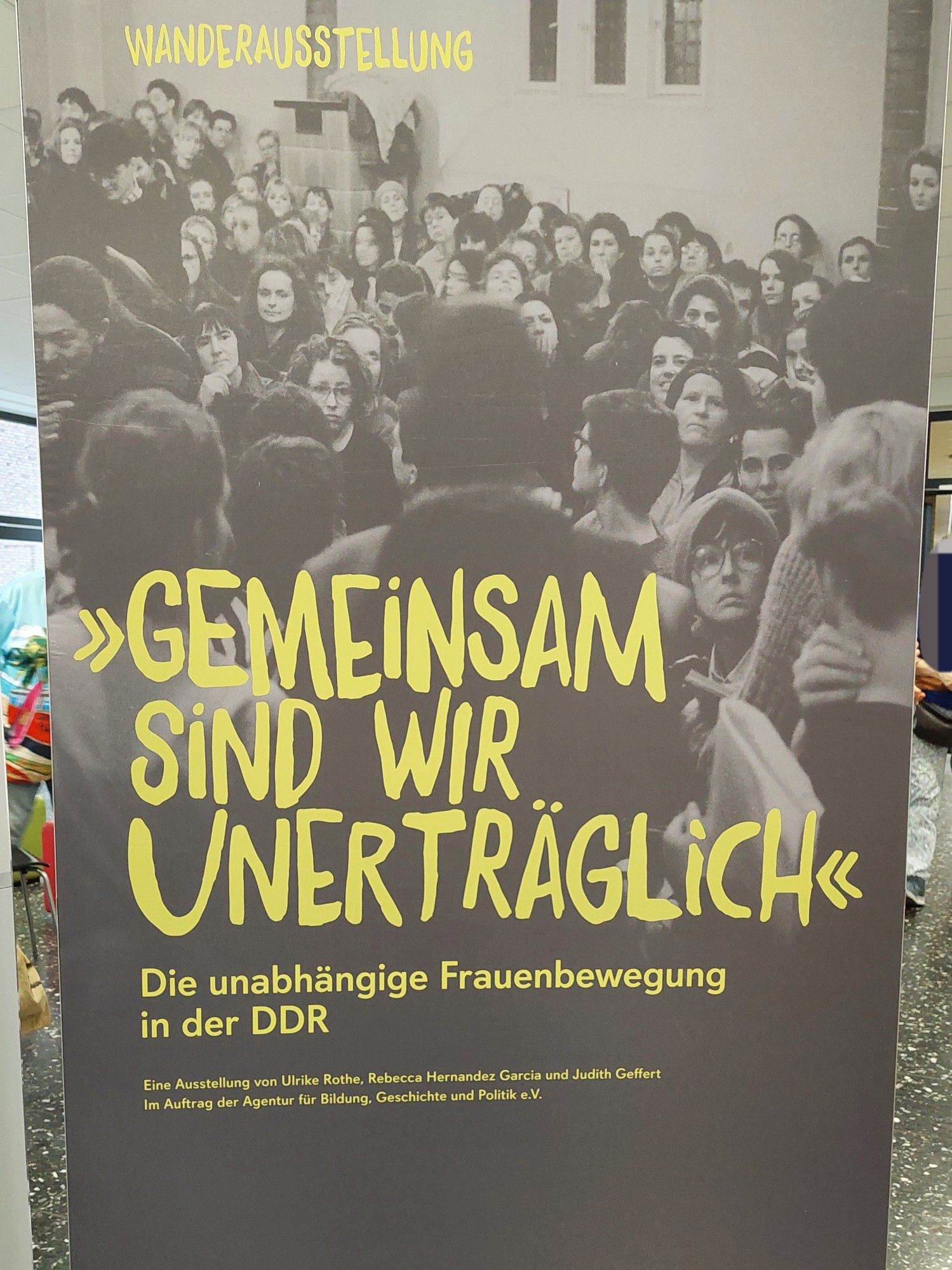 Poster der Ausstellung. Schriftzug "Gemeinsam sind wir unerträglich" auf einem Photo von einem Raum voller Frauen