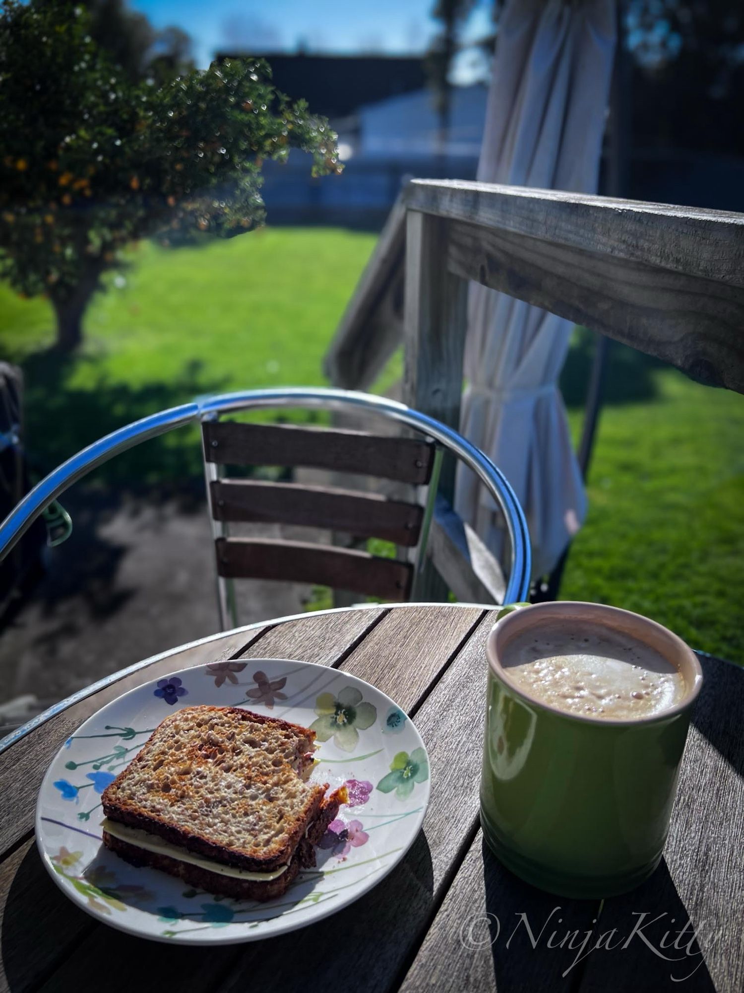 Image by NinjaKitty shows a small wooden table with a toasted sandwich and a large coffee in a green cup on it, outside for the first day of Spring 2024