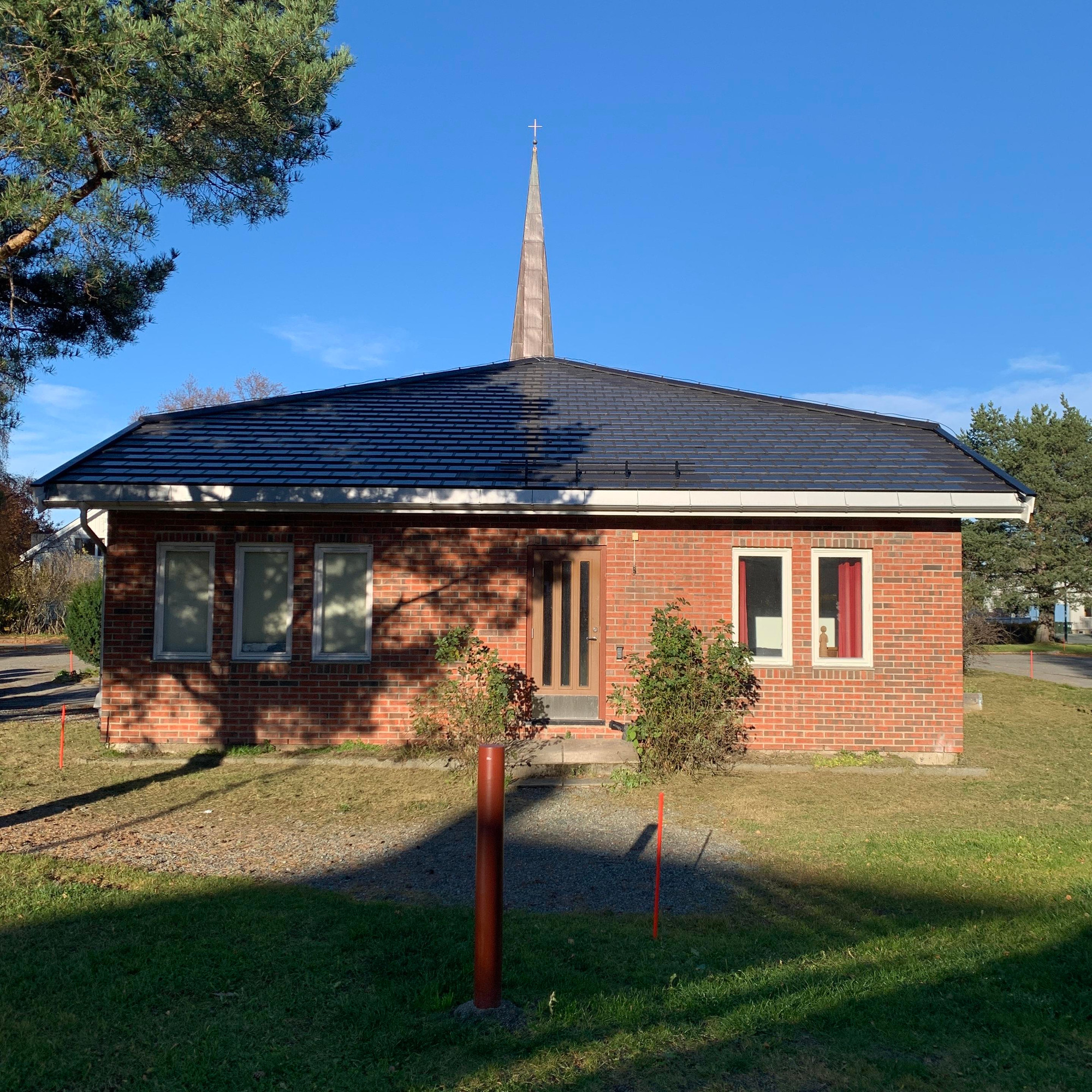 Berg Kirke i sollyset. En furu skygger litt til på venstre side. Kirke har takstein som fungerer som solcellepanel