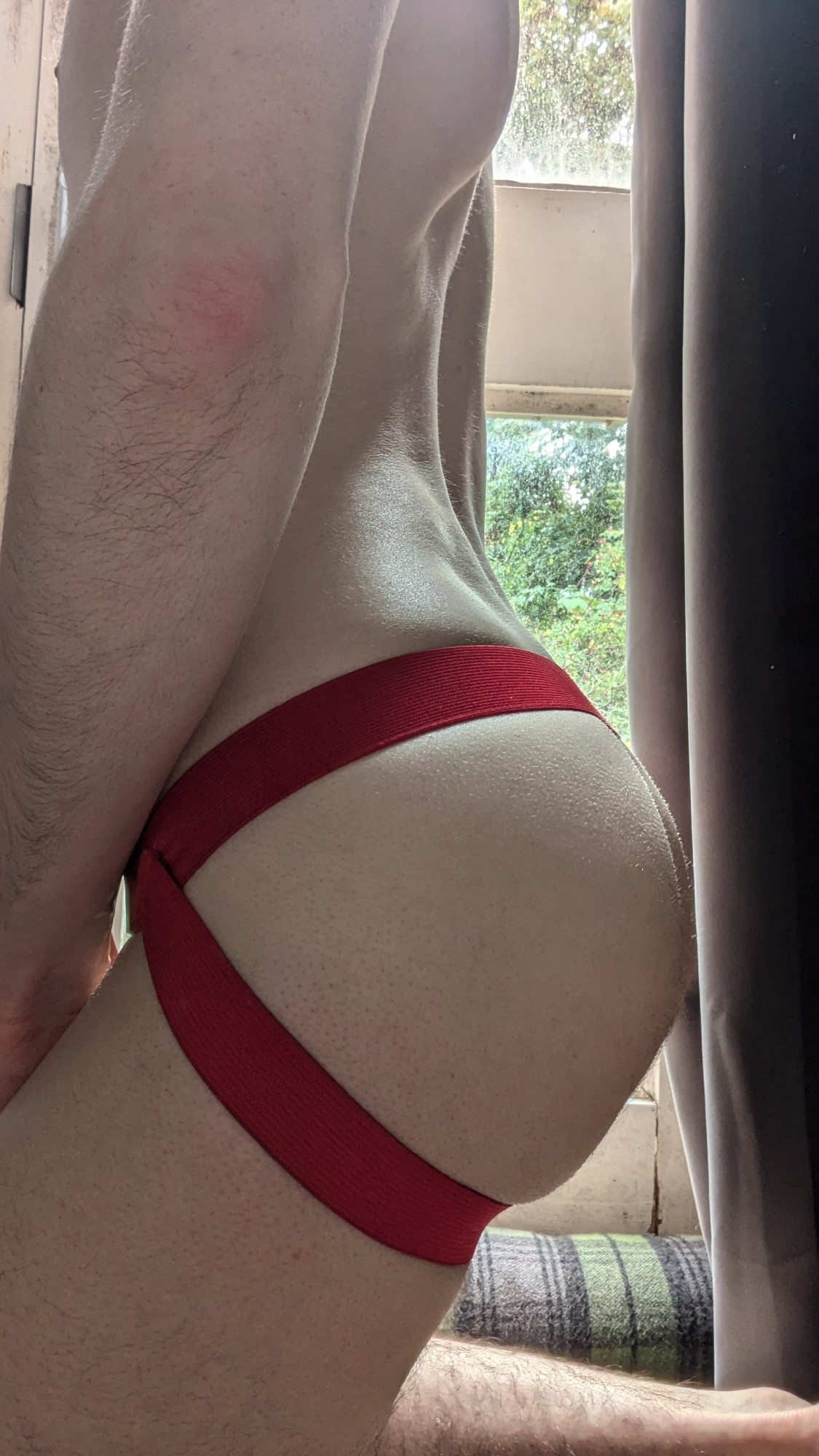 side photo of my pale ass in a bright red jockstrap, kneeling in front of the window