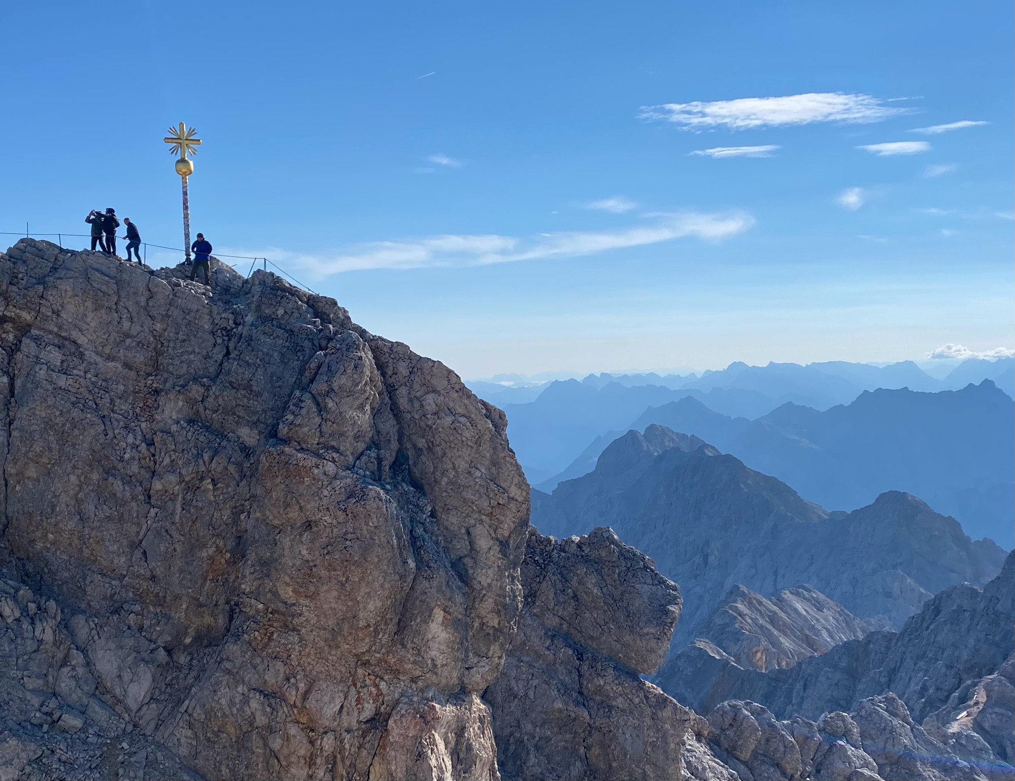 Gipfel der Zugspitze mit Gipfelkreuz, auf der sich vier Personen befinden, dahinter die spektakuläre Berglandschaft