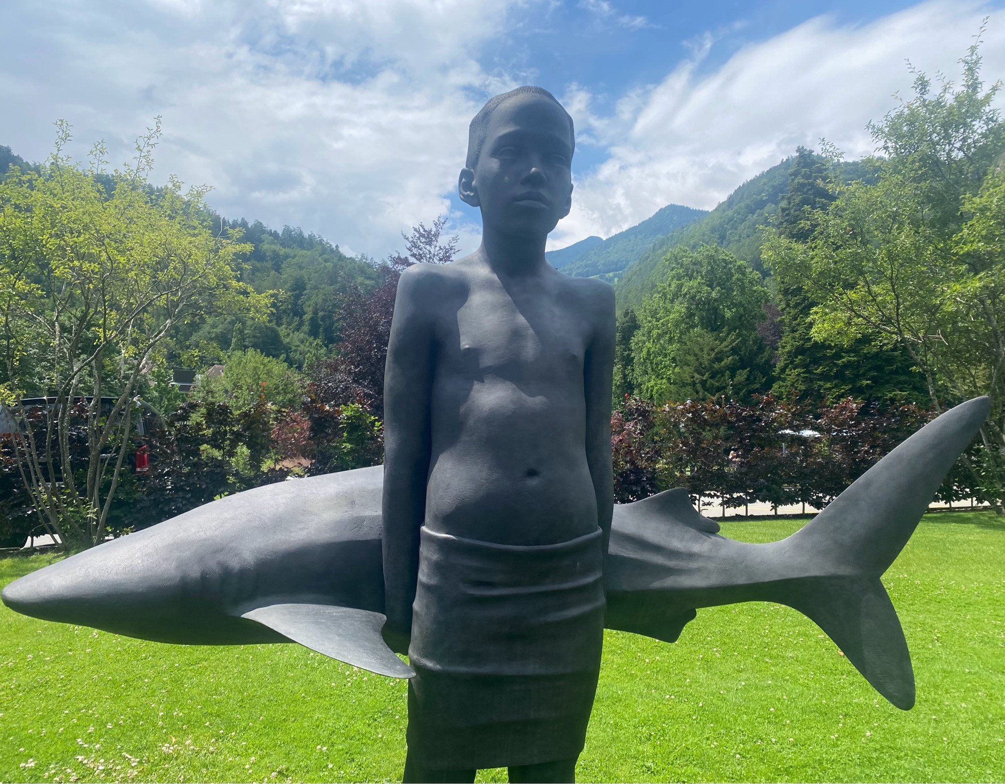 Metallskulptur eines Jungen ab Oberschenkelhöhe, er trägt ein Handtuch wie eine kurze Hose und einen Hai in den Händen hinter dem Rücken. Im Hintergrund Park, Bäume, Berg und bewölkter Himmel.