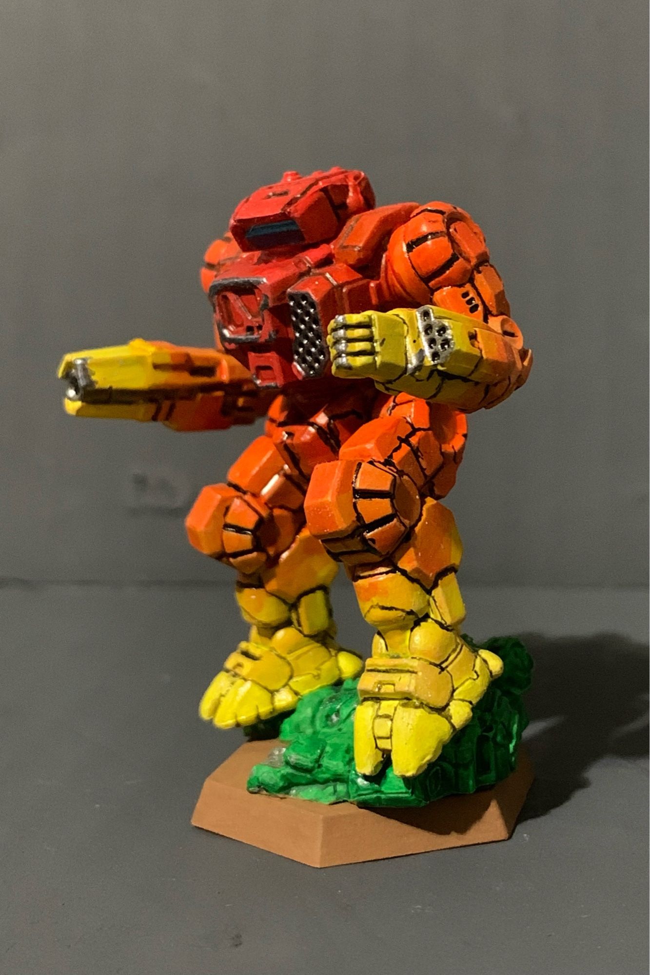 A highlander standing on a crushed and half buried urban mech. It has a red to yellow gradient, going from red in the center torso to yellow on the hands and feet. There is battle damage on the torso, hand, and rifle arm. It is facing slightly to the right.