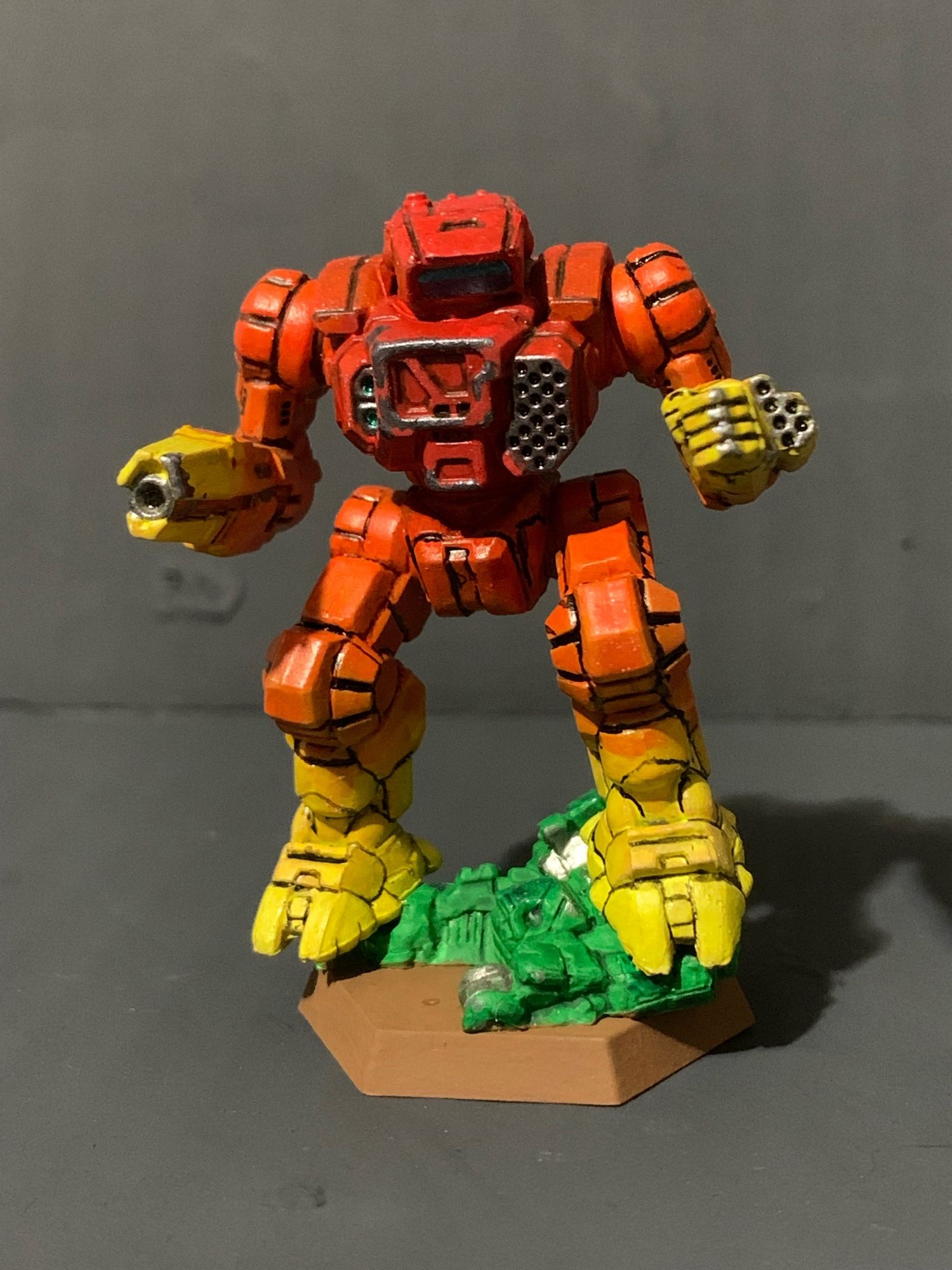 A highlander standing on a crushed and half buried urban mech. It has a red to yellow gradient, going from red in the center torso to yellow on the hands and feet. There is battle damage on the torso, hand, and rifle arm. It is facing forward.