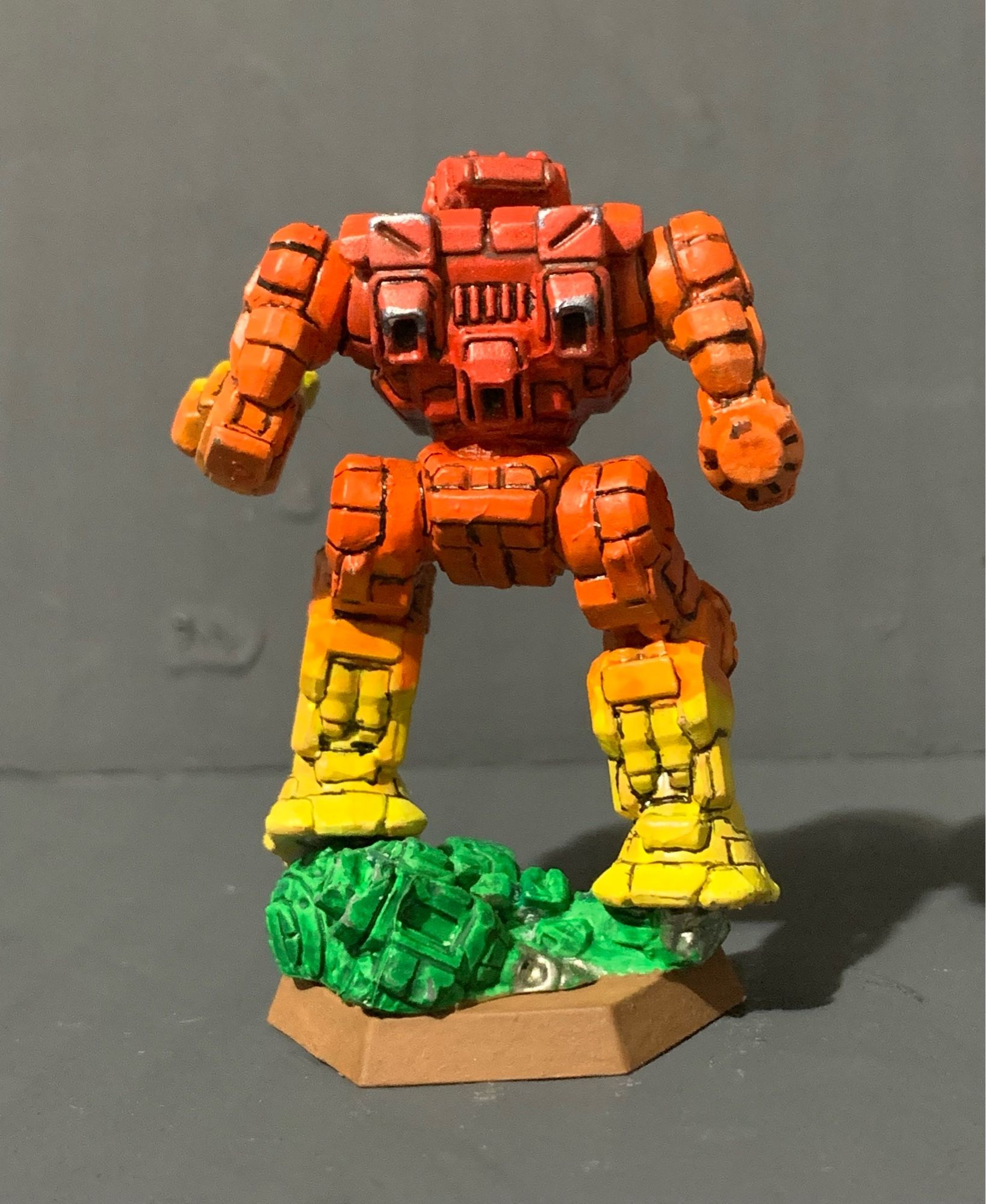 A highlander standing on a crushed and half buried urban mech. It has a red to yellow gradient, going from red in the center torso to yellow on the hands and feet. There is battle damage on its upper back and thruster area. It is facing with its back to the camera.