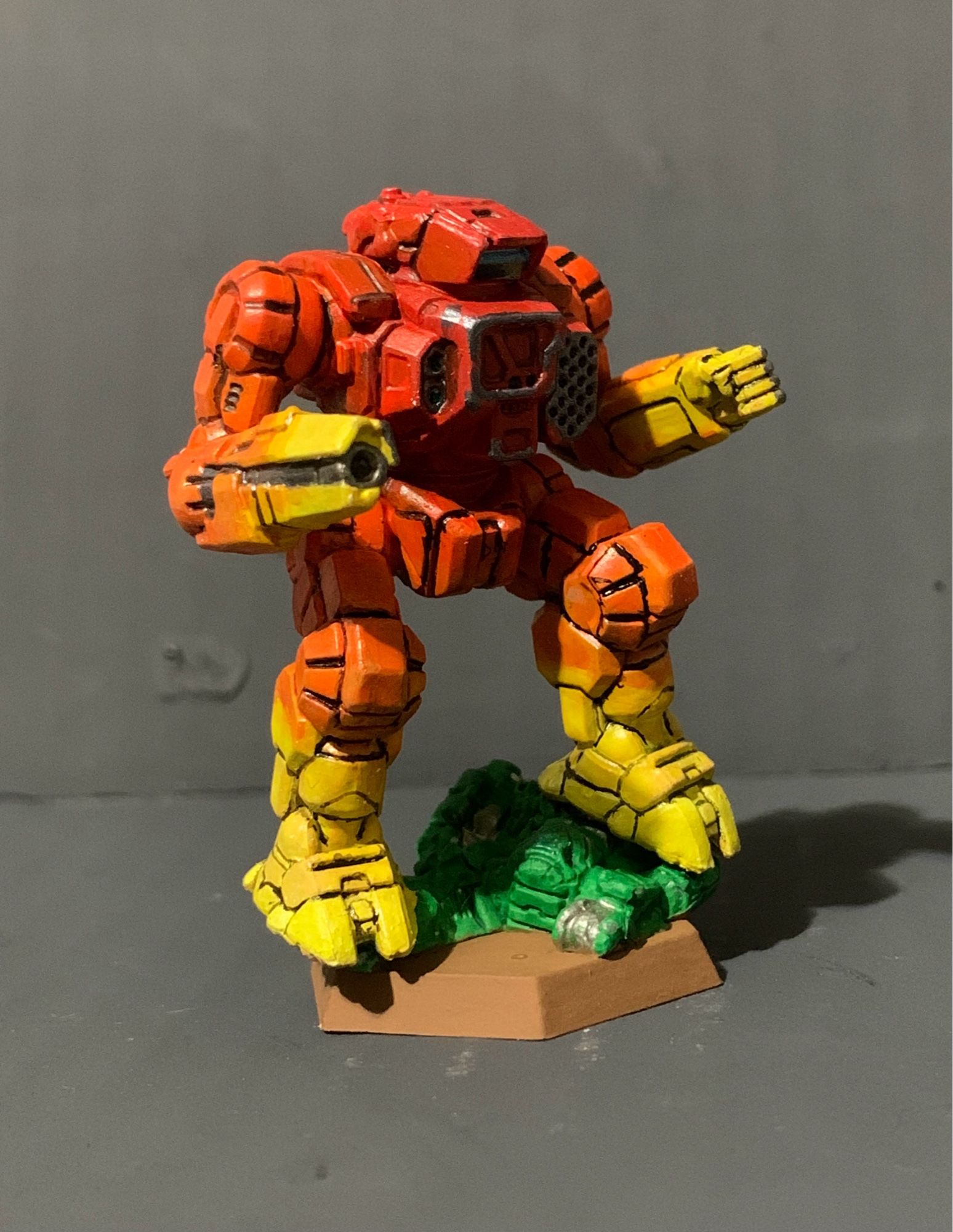 A highlander standing on a crushed and half buried urban mech. It has a red to yellow gradient, going from red in the center torso to yellow on the hands and feet. There is battle damage on the torso, hand, and rifle arm. It is facing slightly left.