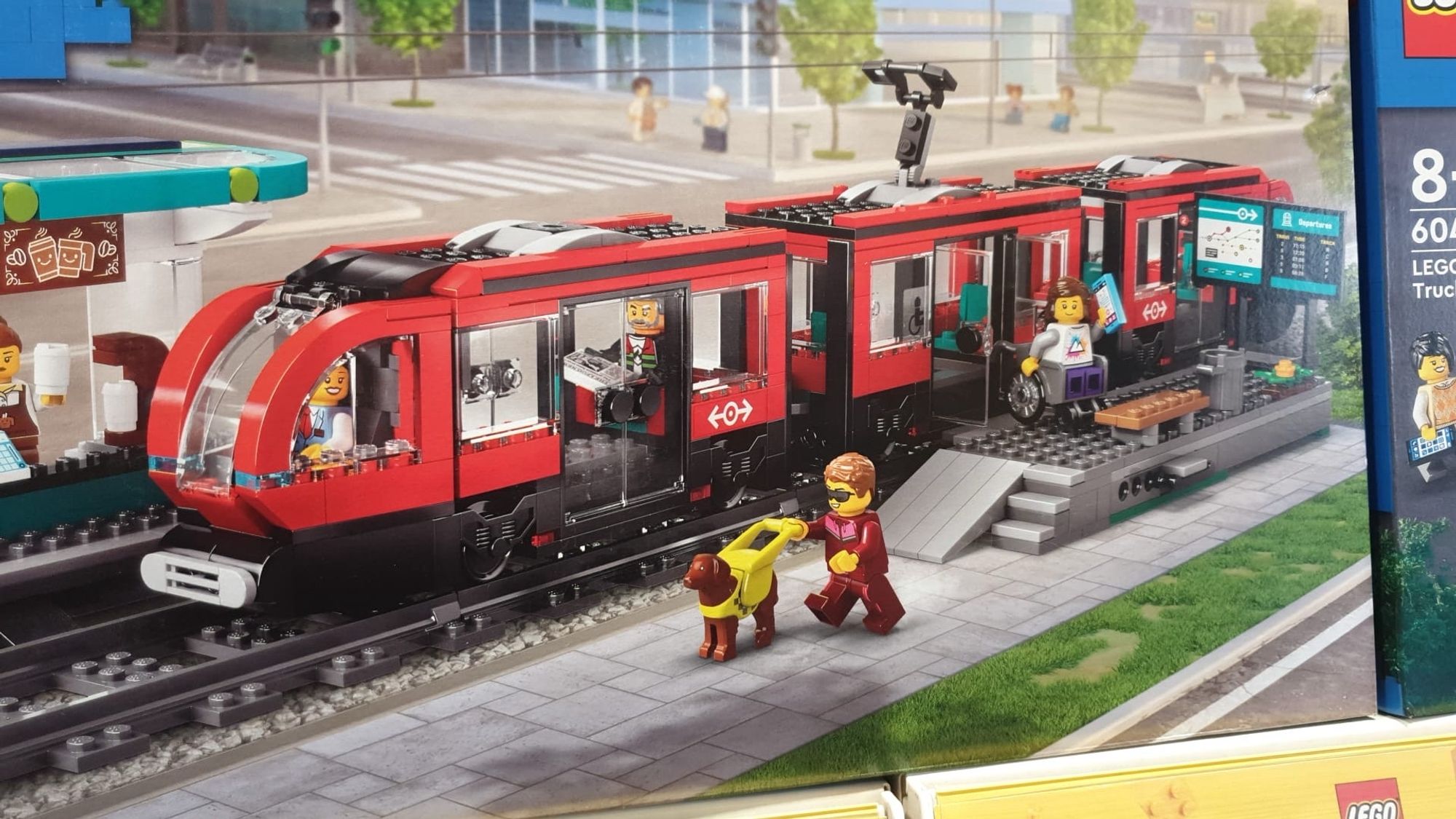 A red Lego train with a raised platform with level boarding and a smiling Lego manual wheelchair user just leaving the train