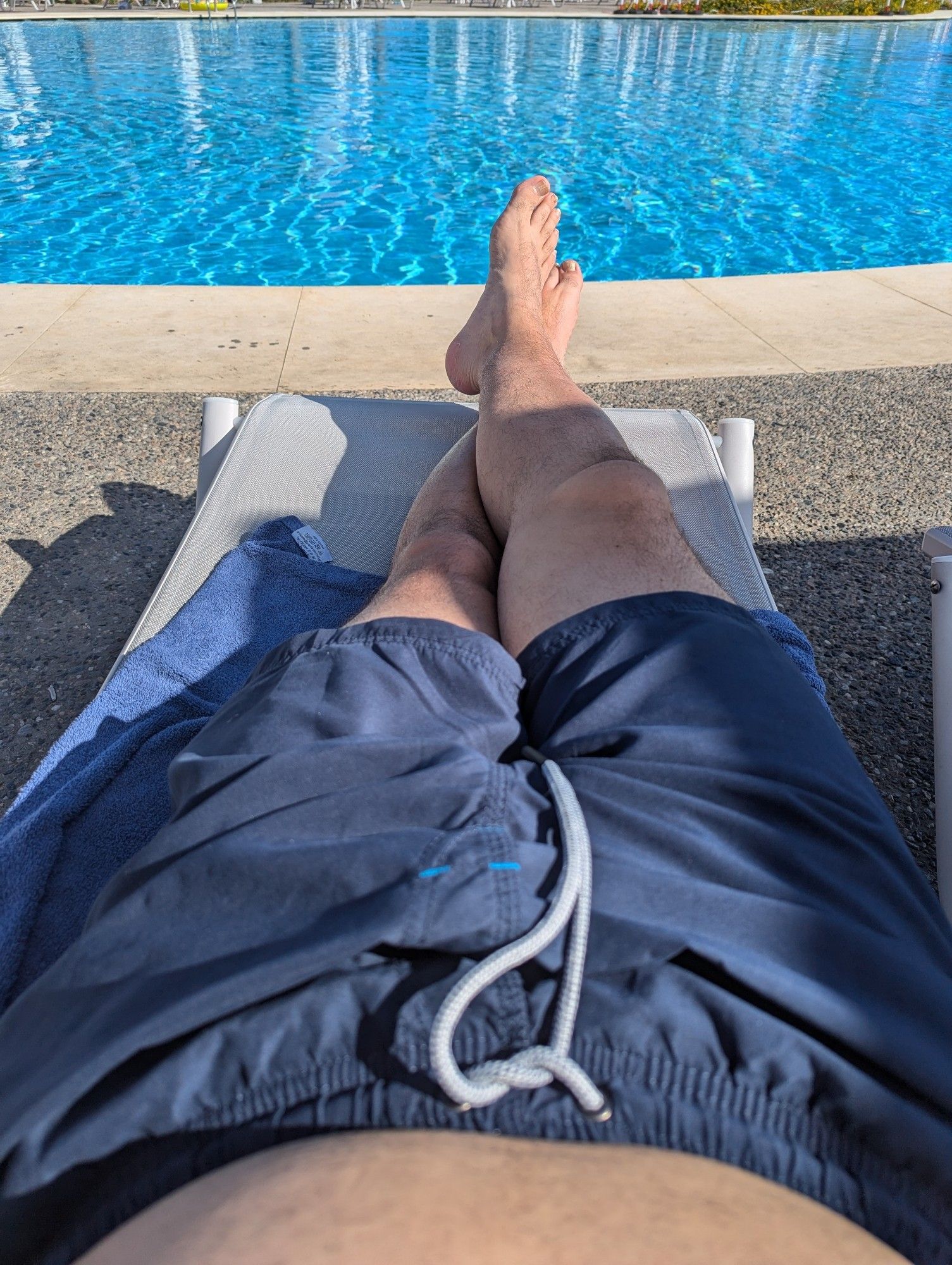 Im Hintergrund blaues Wasser in einem Pool, davor liegt jemand auf einer Sonnenliege, nur die Beine sind zu sehen