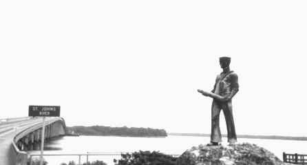 Photo of a bronze statue of a navy guy with a torpedo that looks like a giant dick that is at the base of a bridge in downtown Palatka Florida.