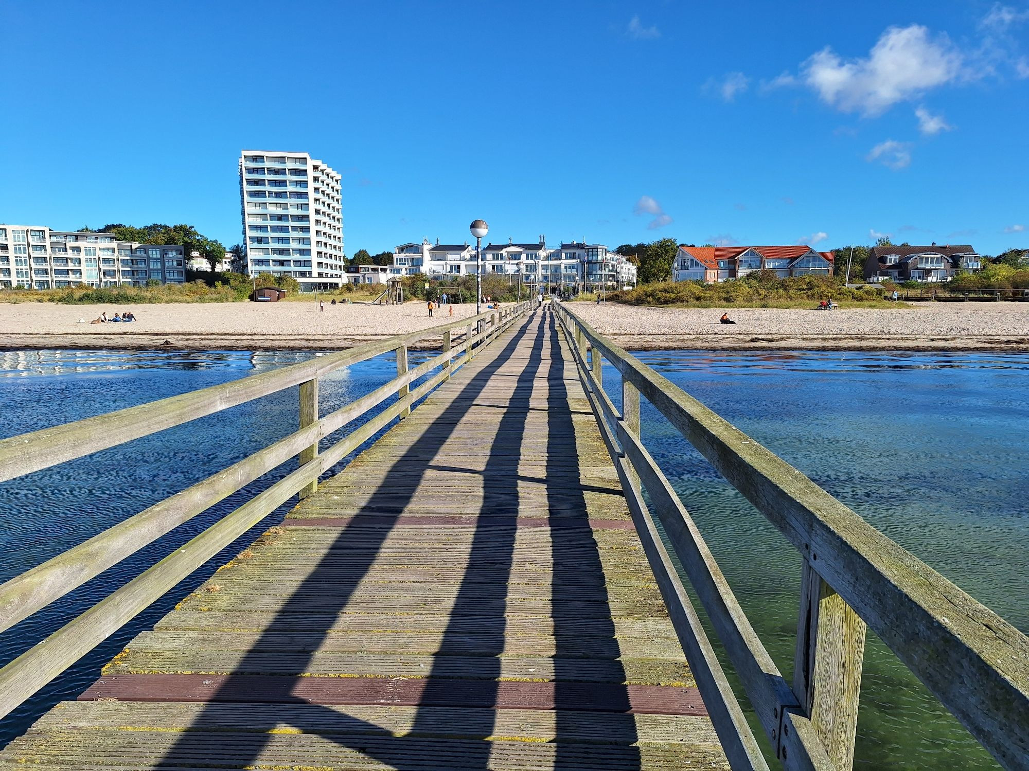 Hölzerne Seebrücke über der blauen Ostsee unter blauem Himmel mit Blick auf einen Ort mit einem weißen Hochhaus.