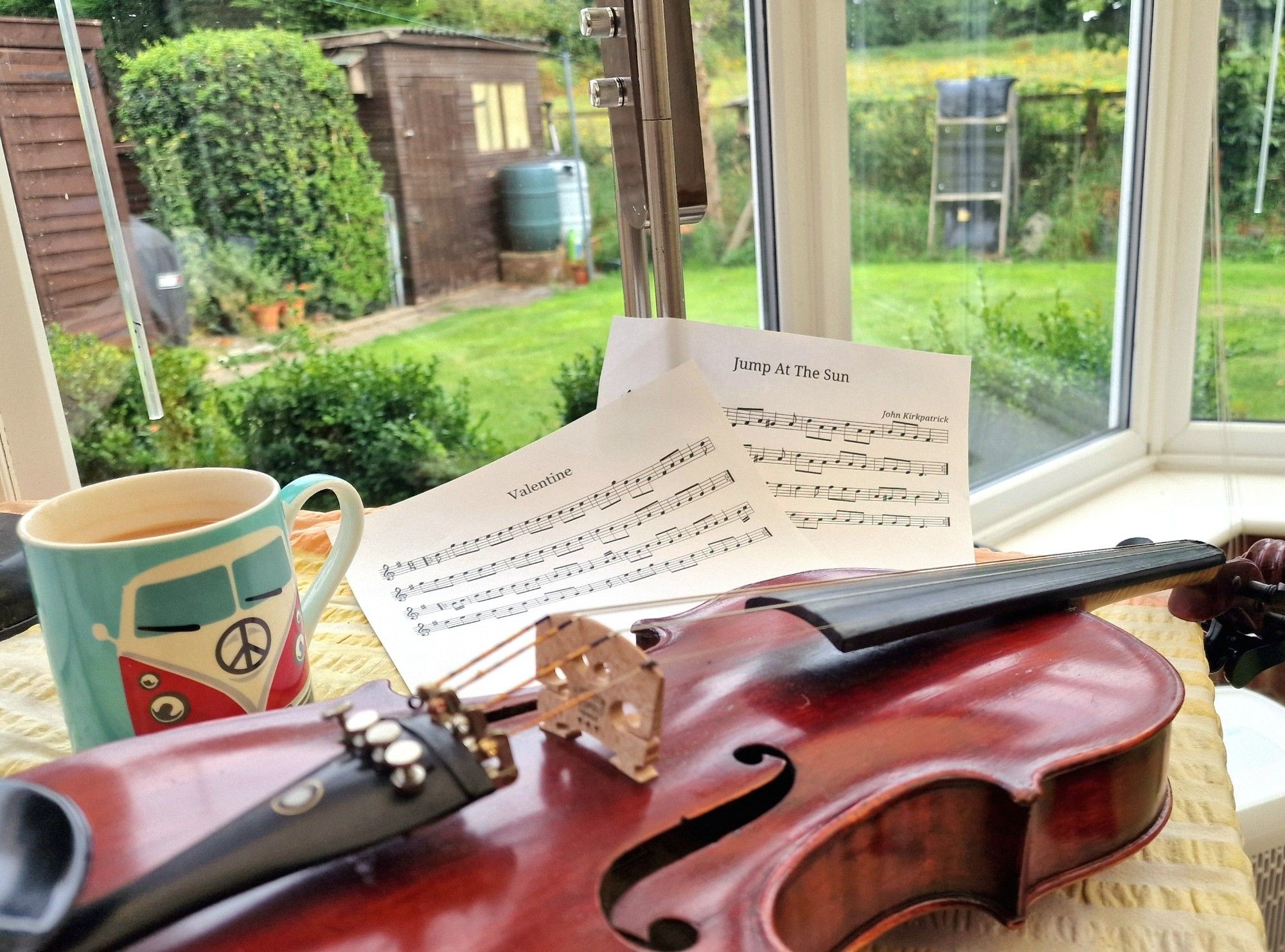 Image shows a violin on a table with 2 sheets of music. Through a window a garden is visible.