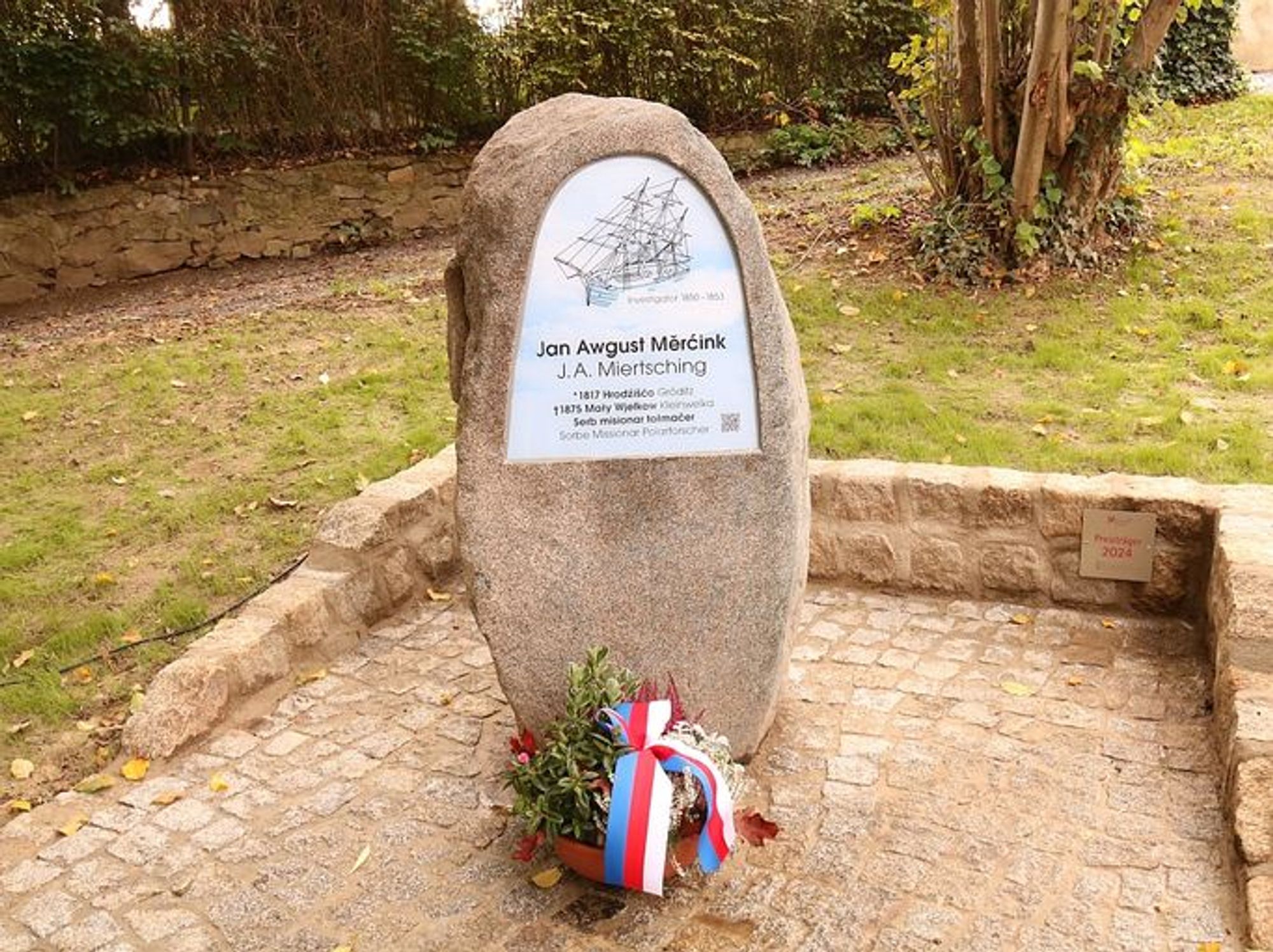 Das Bild zeigt ein Denkmal - einen Findling, an den eine Tafel montiert ist, die oben die Silhouette eines Schiffs zeigt, darunter Namen und Daten der geehrten Person Johann August Miertsching in sorbischer und deutscher Sprache