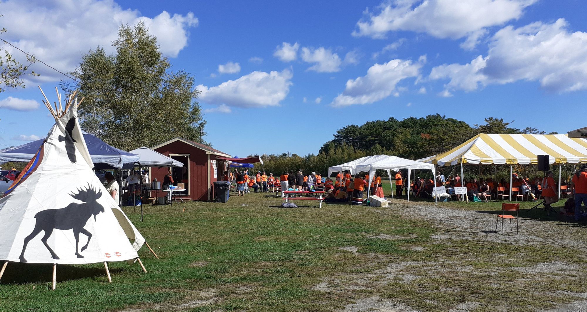 Das Bild zeigt eine Wiese mit einem Tipi und Festzelten sowie vielen Frauen, Männer und Kinder. Ein großer Teil von ihnen trägt ein orangenes T-Shirt.