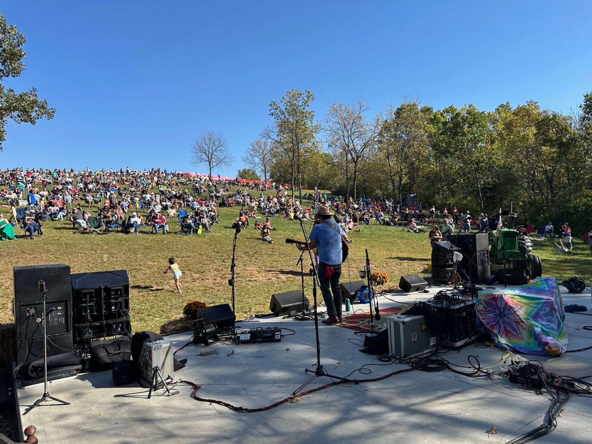 POV from behind stage showing a man playing guitar with a crowd on the expanse of the hillside.