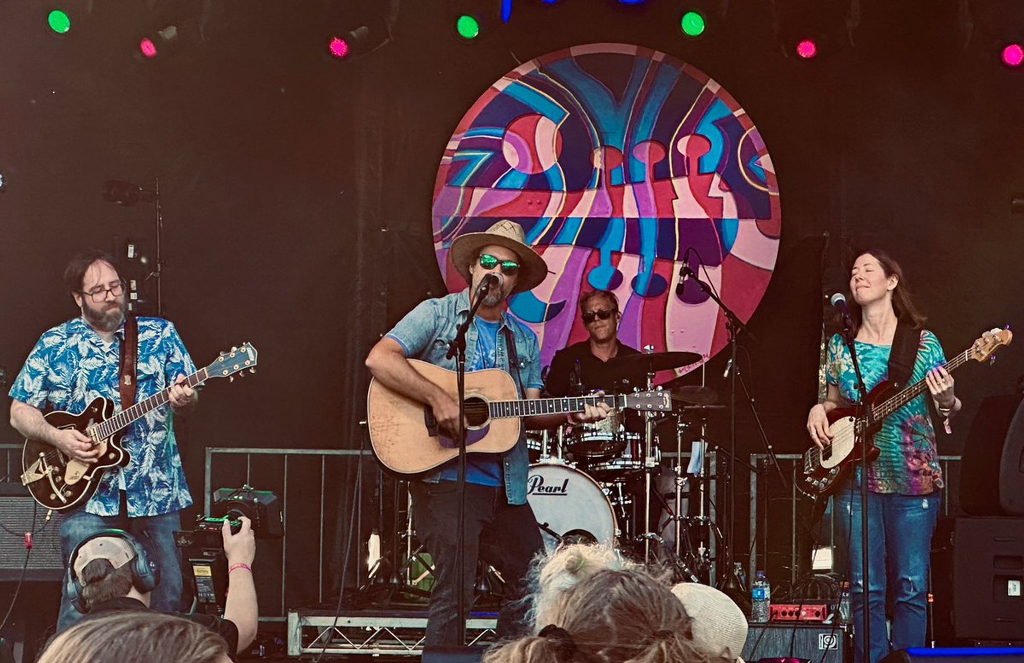 Four musicians on stage.  From left; man playing guitar, man playing guitar and singing, man playing drums, woman playing electric bass.