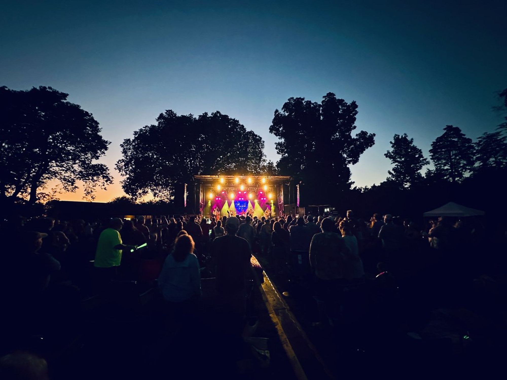 The sun is setting behind the stage, which is surrounded by trees.  The stage is lit in red, blue and yellow.