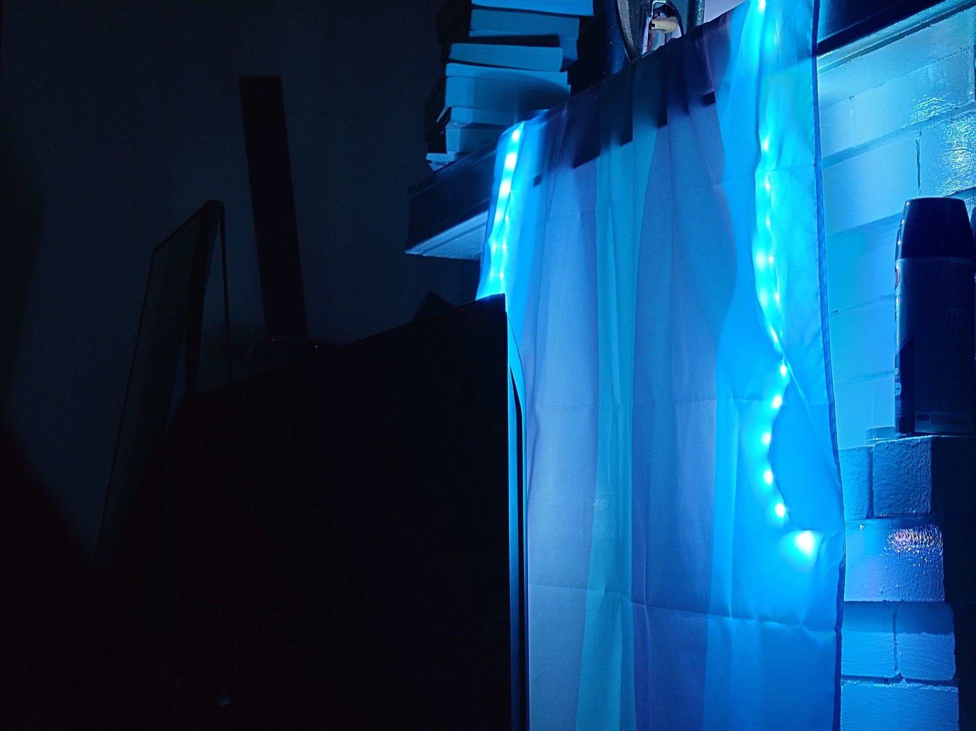 photo of dark room lit solely by the blue LED strip under a trans pride flag, hanging on a white, stone mantle alongside books, a skateboard, and a can of spray paint