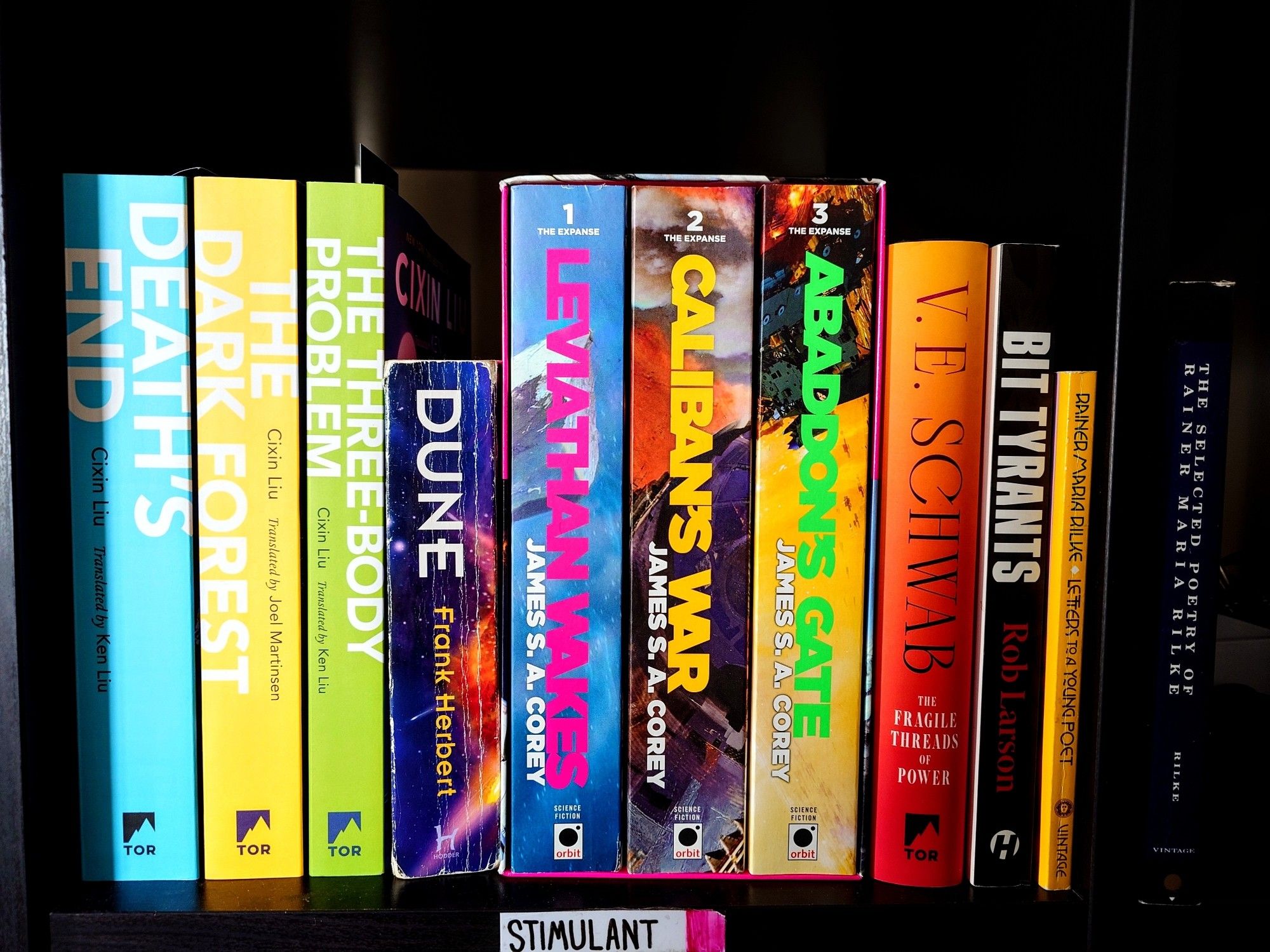 a row of colorful books on a black shelf, in order (by name of series/book from left to right): The Three Body Problem Series, Dune, The Expanse Trilogy, The Fragile Threads of Power, Bit Tyrants, Rilke's Letters To A Young Poet, and The Selected Poetry Of Rainer Maria Rilke