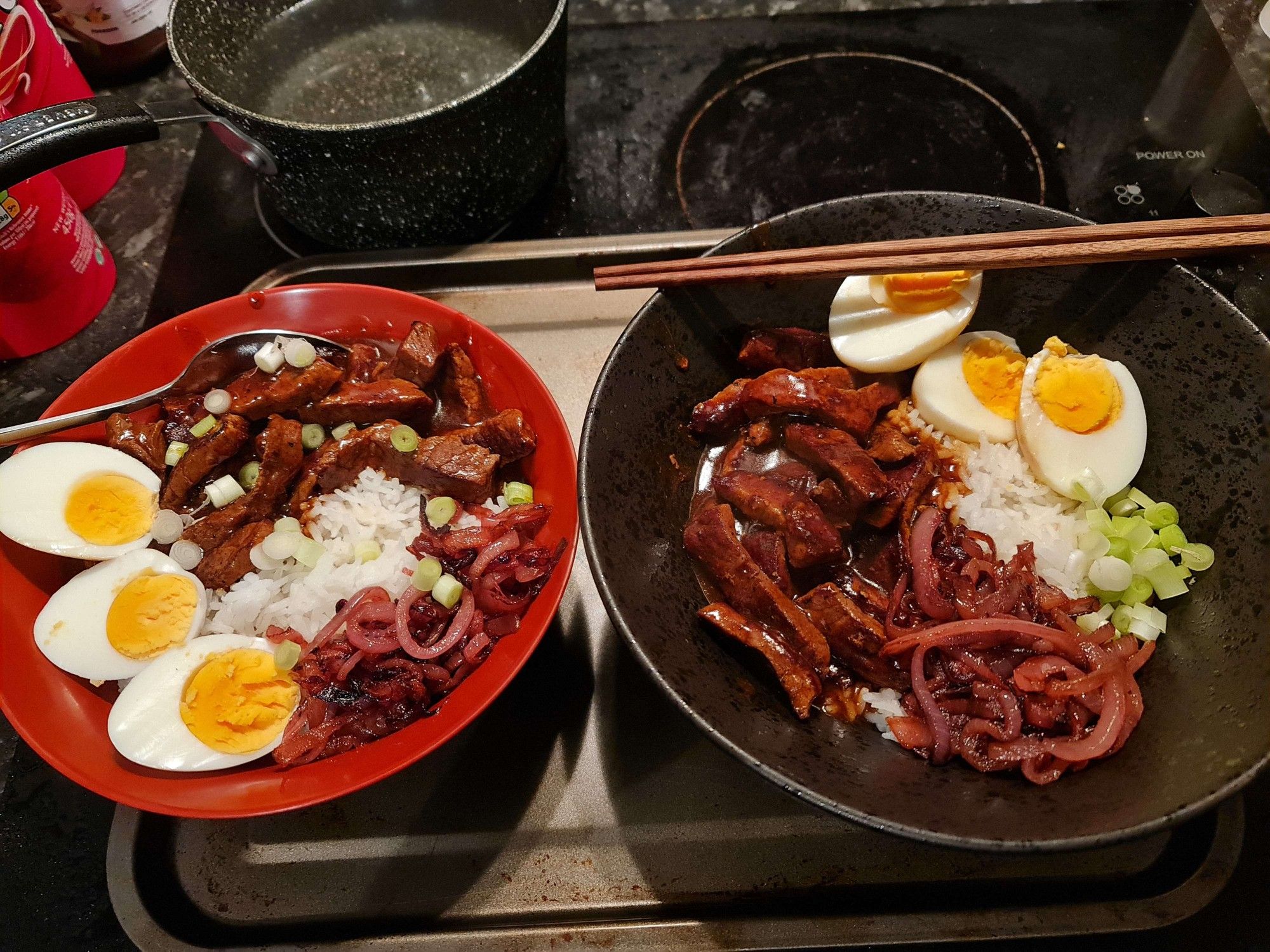 Two bowls with White Rice, Strips of Beef and Caramelized Onions inside. 3 halves of Egg lie on top and there are various Spring Onions scattered as a topping. The left bowl is a Red Ramen Bowl and has a spoon inside. The right bowl is a bigger ceramic black Ramen Bowl and has chopsticks on top.