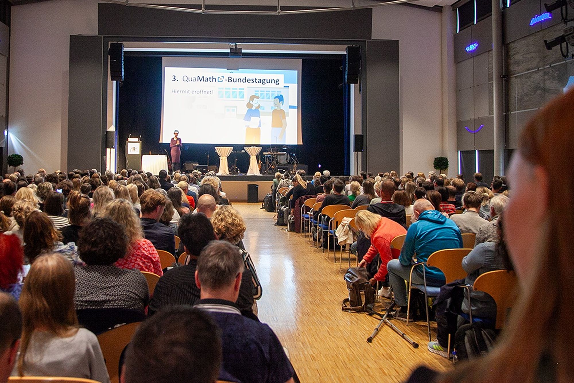 Auftakt zu 3. QuaMath-Bundestagung im Stadtsaal Dillingen. Blick durch den vollbesetzten Saal auf eine Leinwand mit Rednerin auf der Bühne.