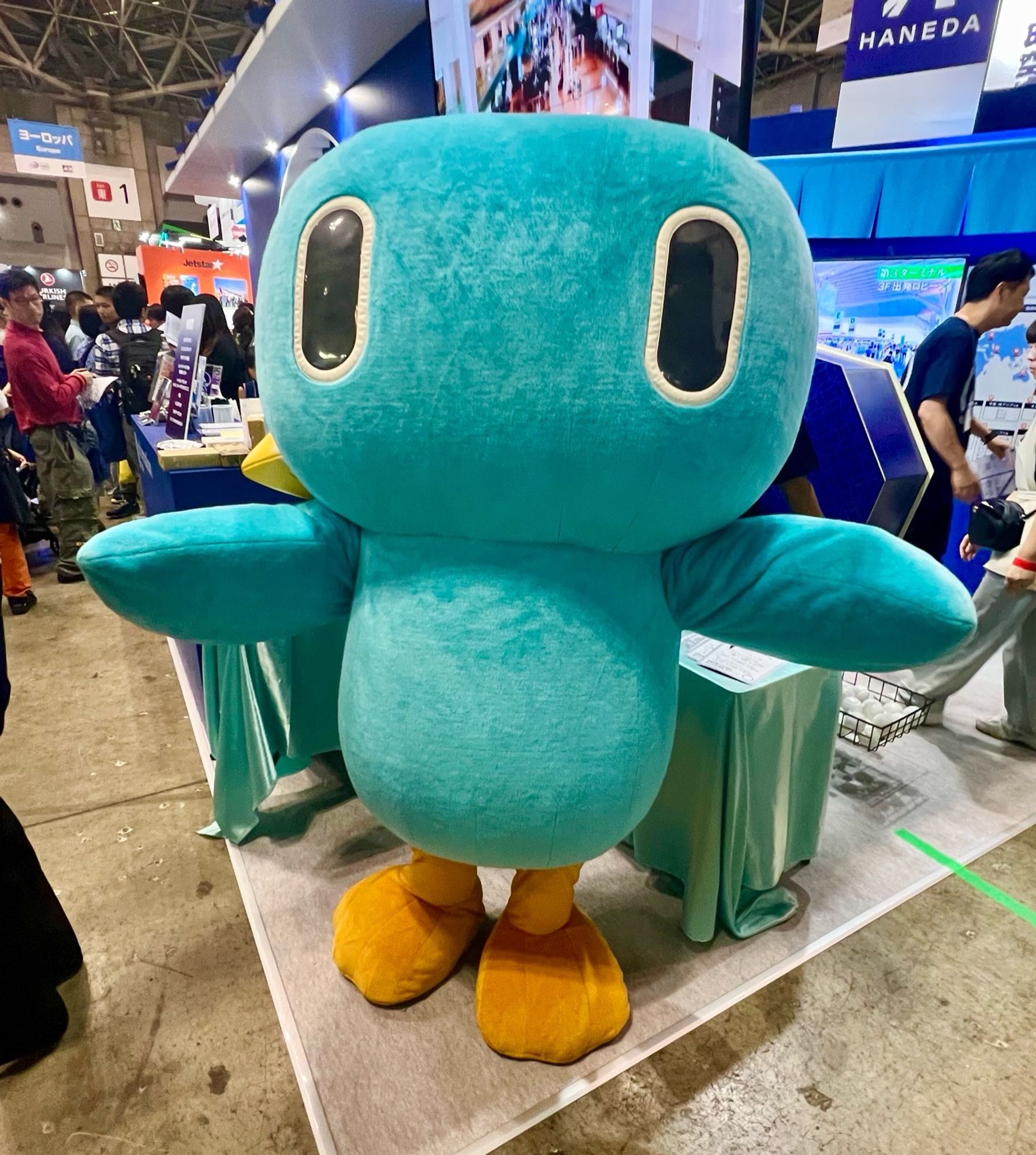 A bluebird mascot whose beak is on the side of its head spreads its wings in a crowded convention centre.