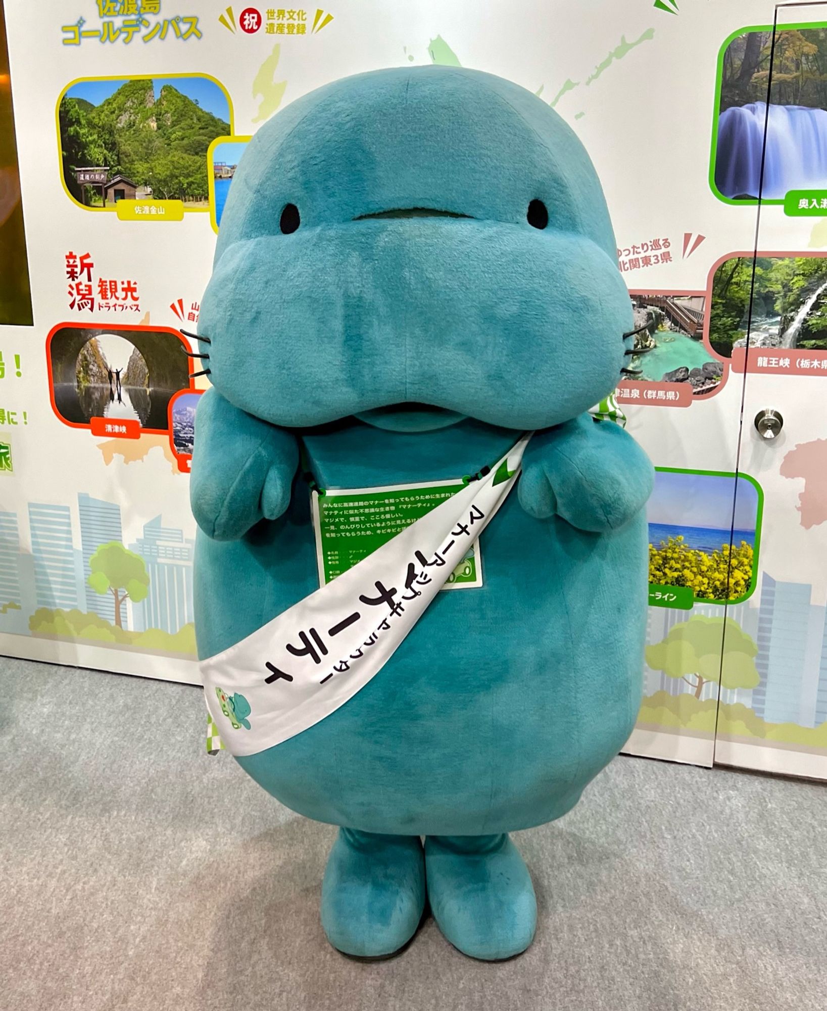 A blue manatee mascot with legs stands in front of a wall featuring landscape photos of tourist attractions.