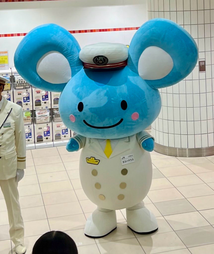 A blue mouse with no nose is standing on two legs and wearing a white stationmaster’s uniform.