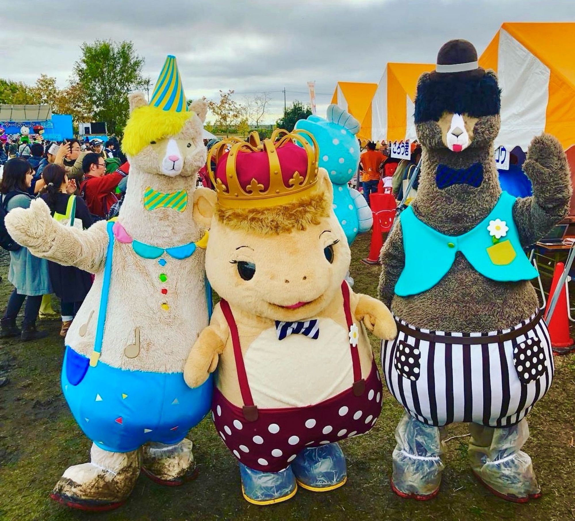 Three llama mascots wearing hats and pants with suspenders stand in a crowded field.