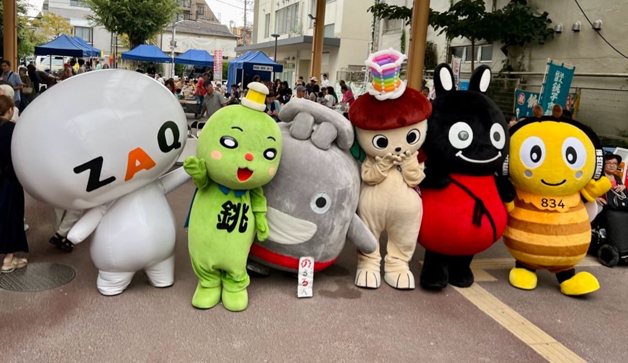 Six regional mascots stand in a row in a crowded town square.